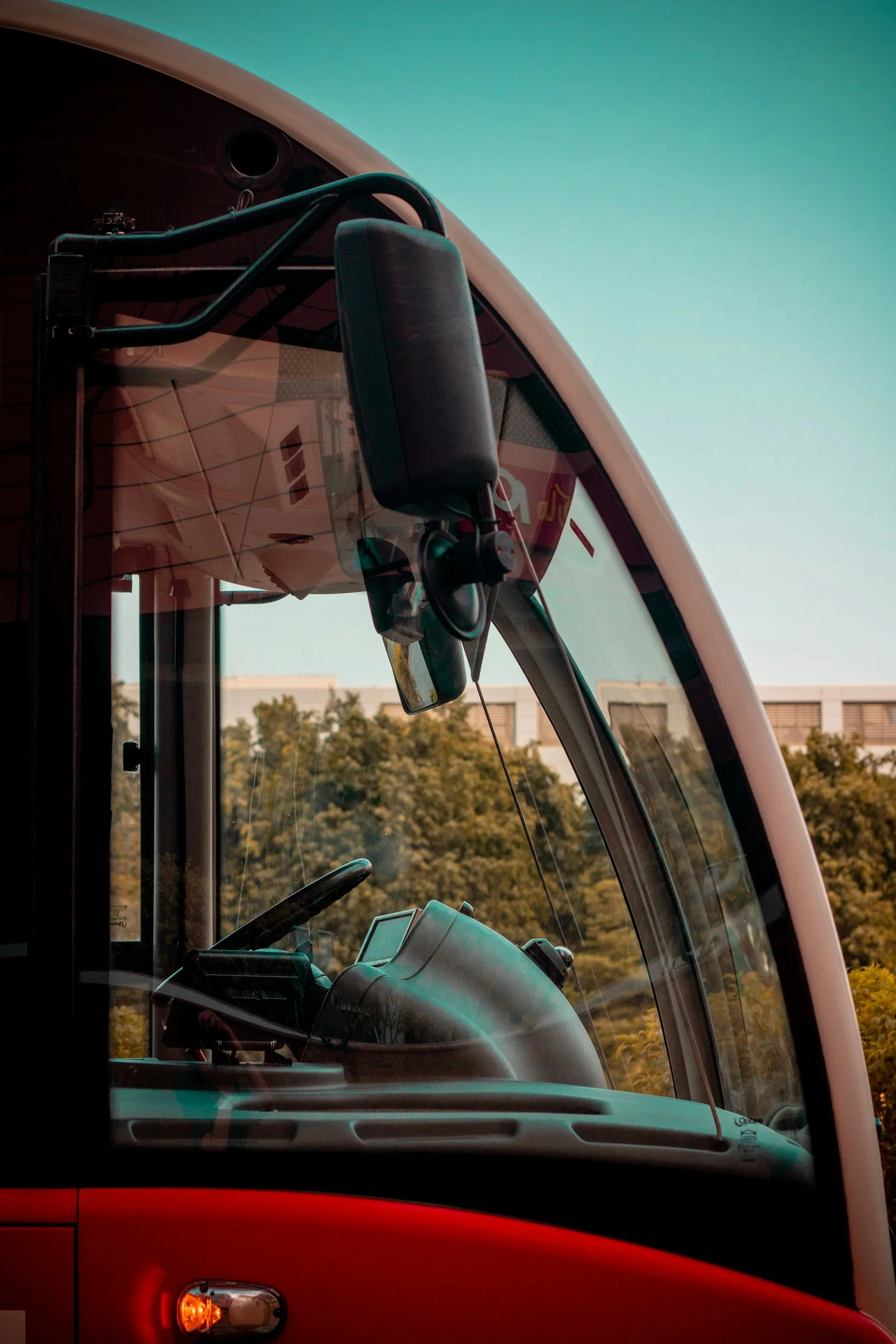 Close-up of the front cabin area of a modern truck or bus with a large curved windshield, side mirror, and interior dashboard, set against a backdrop of trees and a building.
