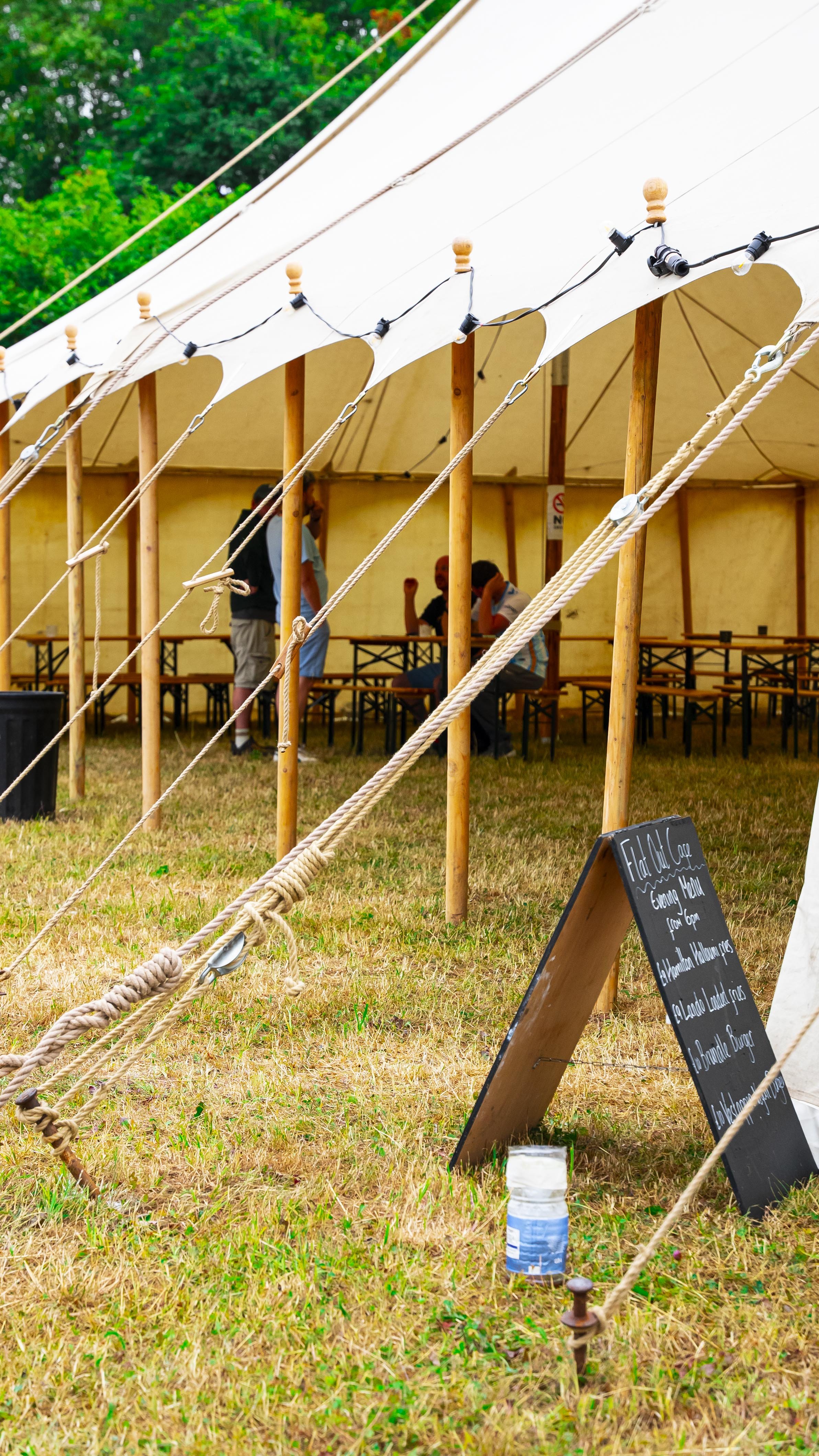 Inside a large yellow tent, there are tables and chairs with people sitting and standing around. The tent is supported by wooden poles and secured with thick ropes. There is a signboard on the grass in front of the tent.