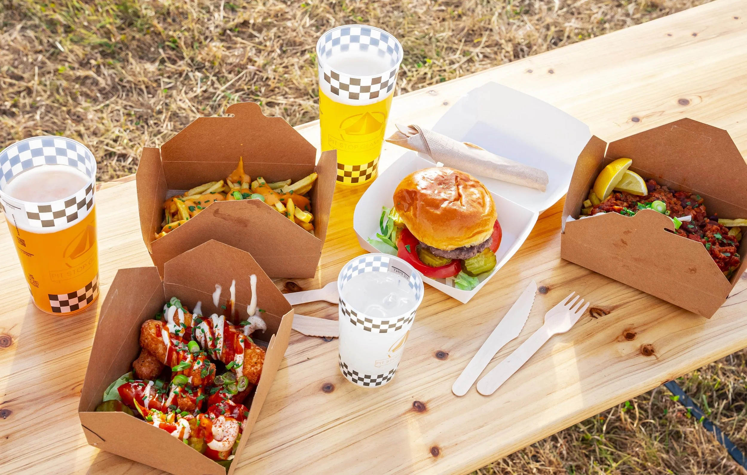 Outdoors meal with burgers, fries, chicken bites, and drinks on a wooden table.
