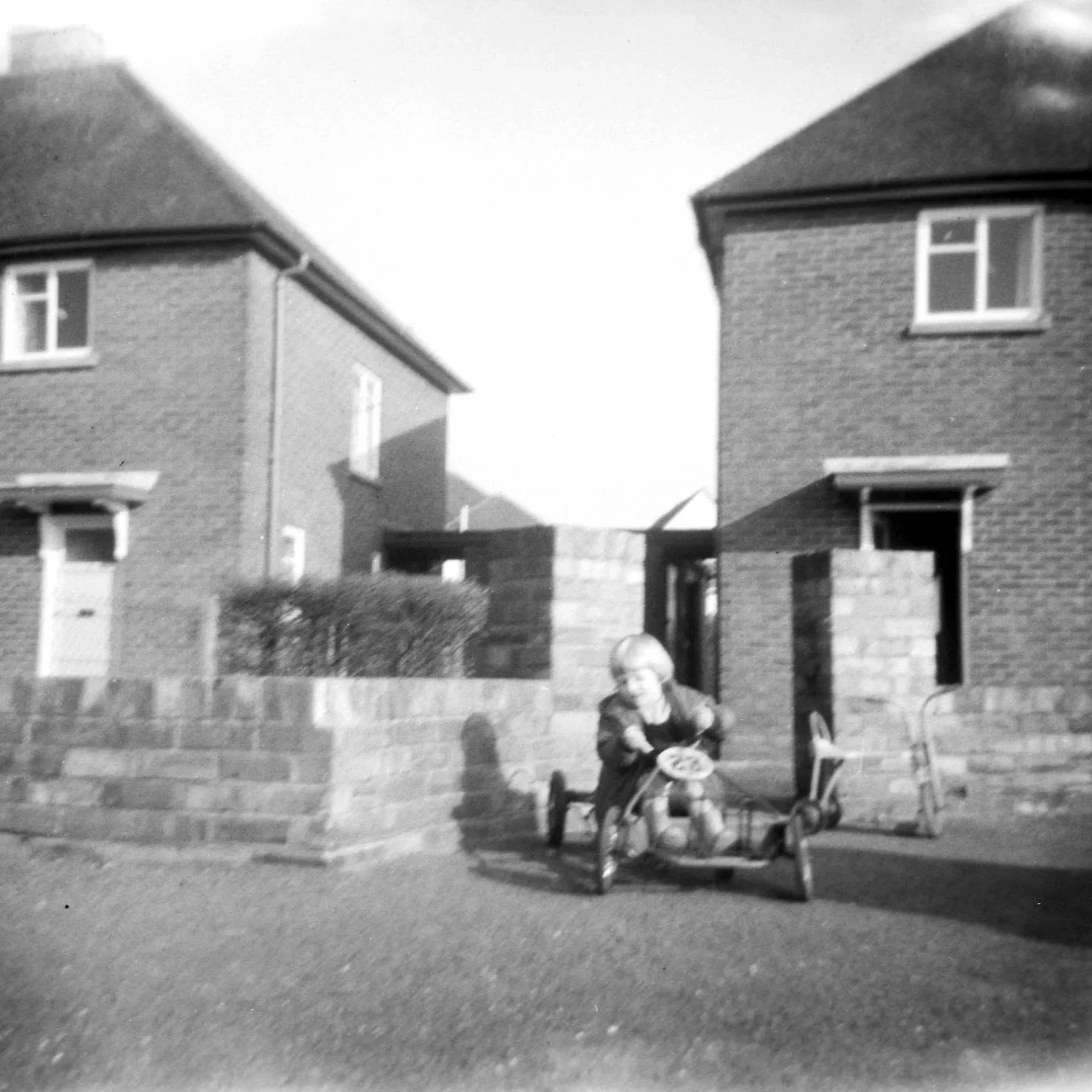 A black and white photo of a young girl riding a tricycle in front of a brick wall, with two houses in the background. The girl appears to be holding a book or magazine, and there is a small chair and a doll or stuffed animal nearby.