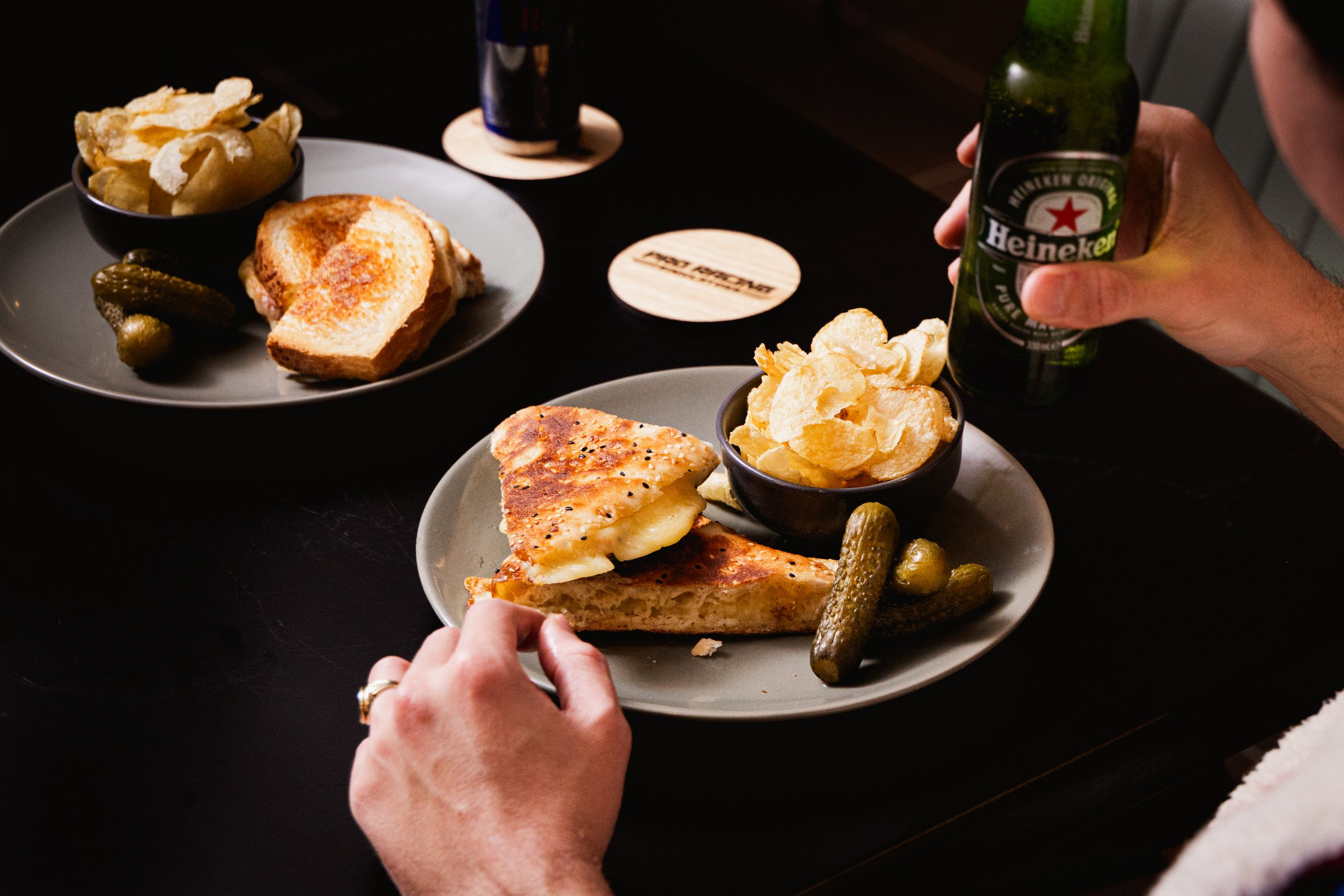 People sharing plates of food including grilled cheese sandwiches, potato chips, pickles, and possibly a chicken dish, with someone holding a Heineken beer bottle in the foreground.