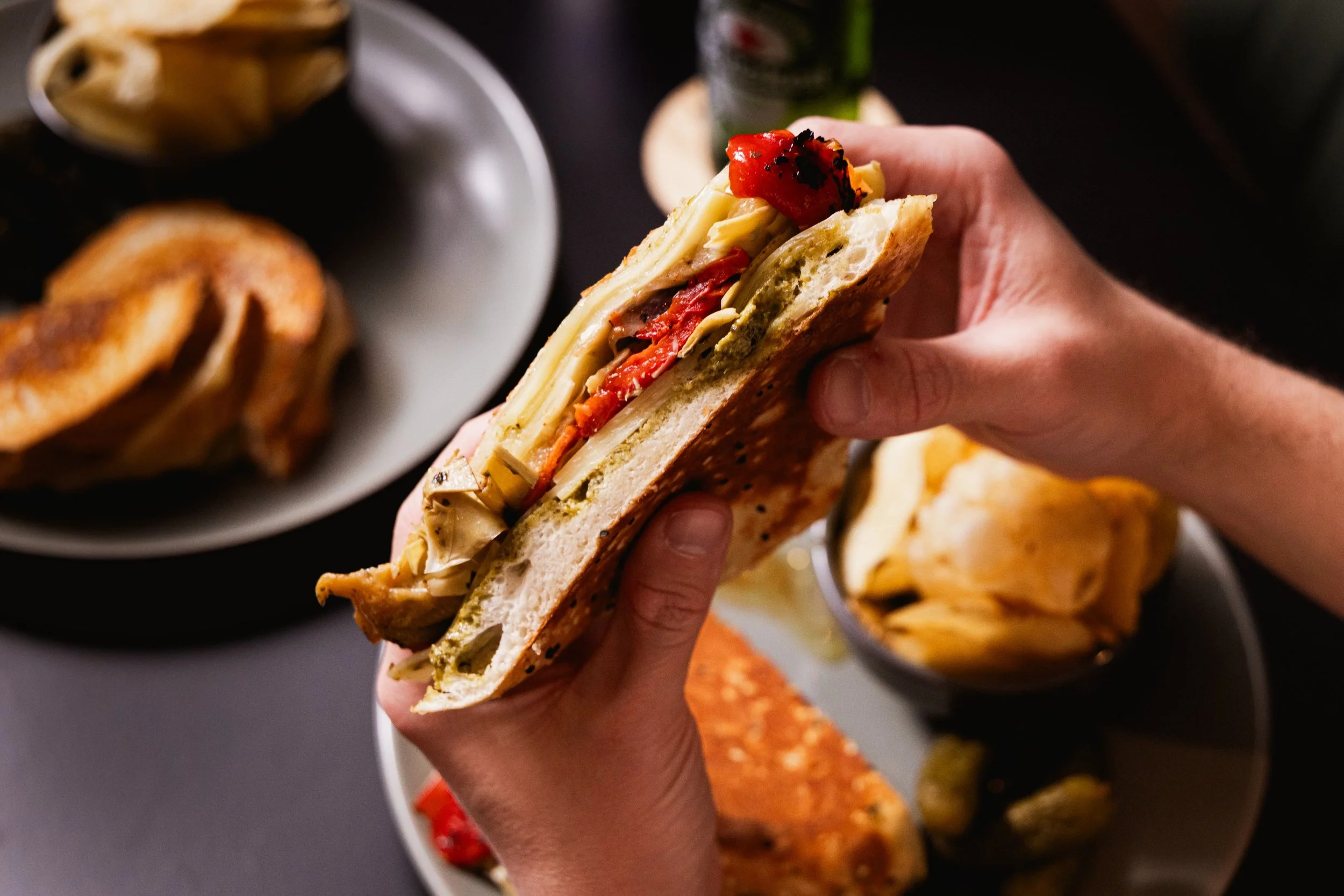 Hand holding a sandwich with cheese, red pepper, and sliced bread. In the background, there are plates with potato chips, a grilled burger bun, and salsa or nachos.