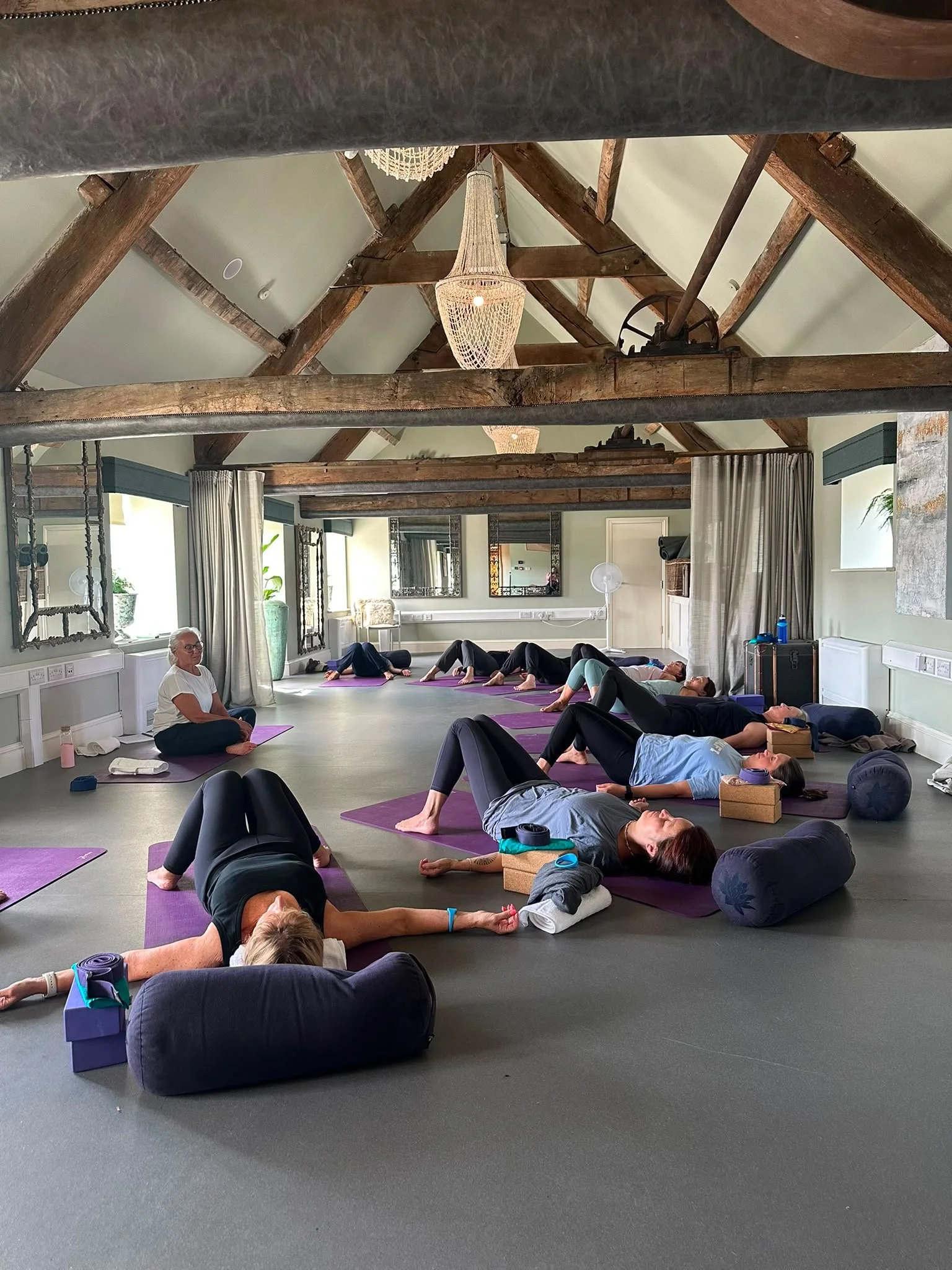 A group of people participating in a yoga class in a spacious room with wooden beams and large windows, lying on purple yoga mats and relaxing.