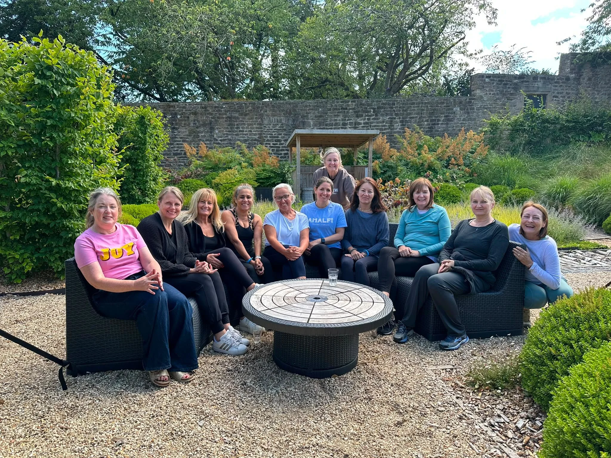 A group of twelve women sitting outdoors in a garden with lush green plants and trees behind them, sitting around a round table.