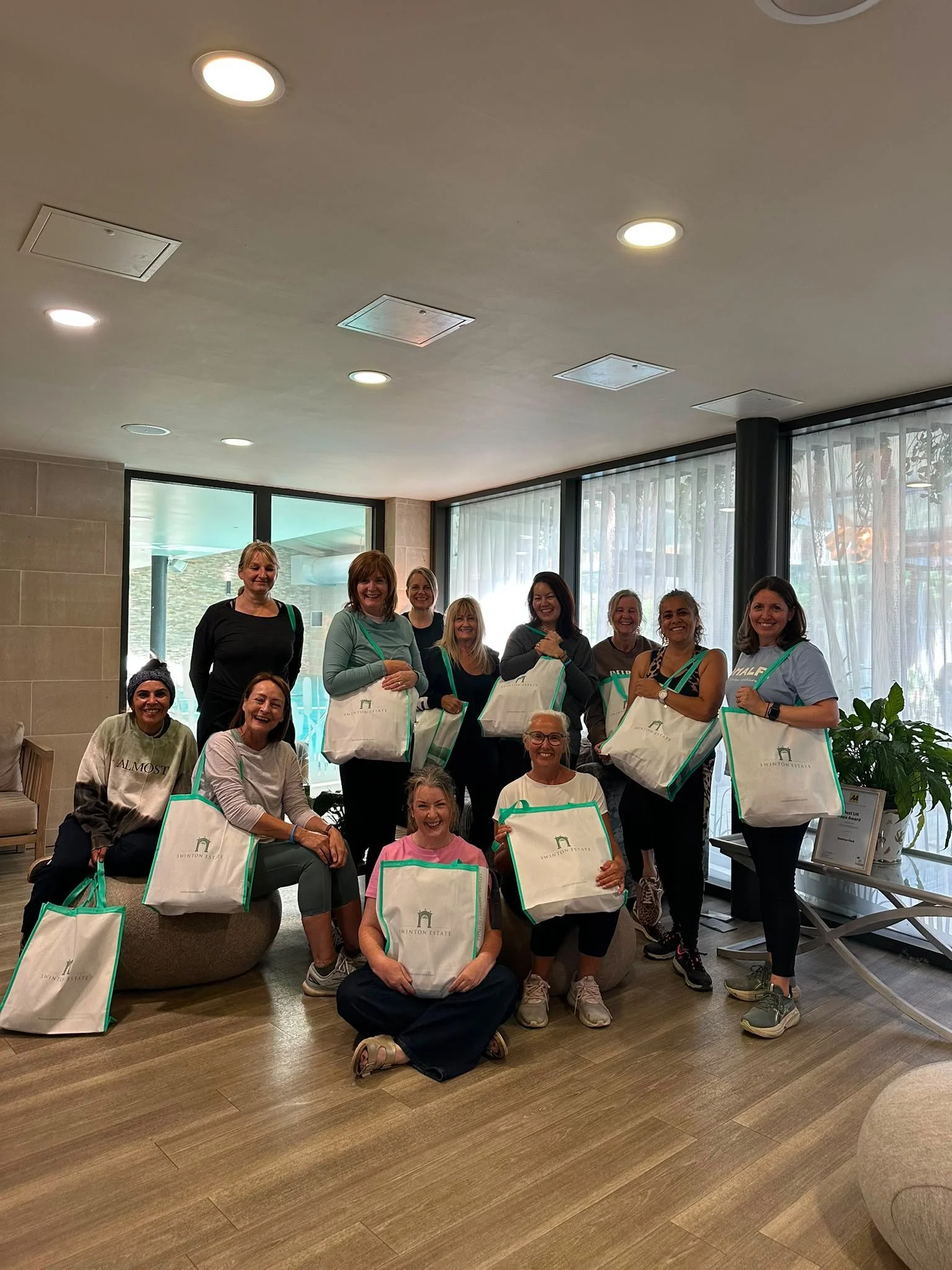 A group of women posing with tote bags in a modern indoor space with large windows and curtains, celebrating or attending an event.