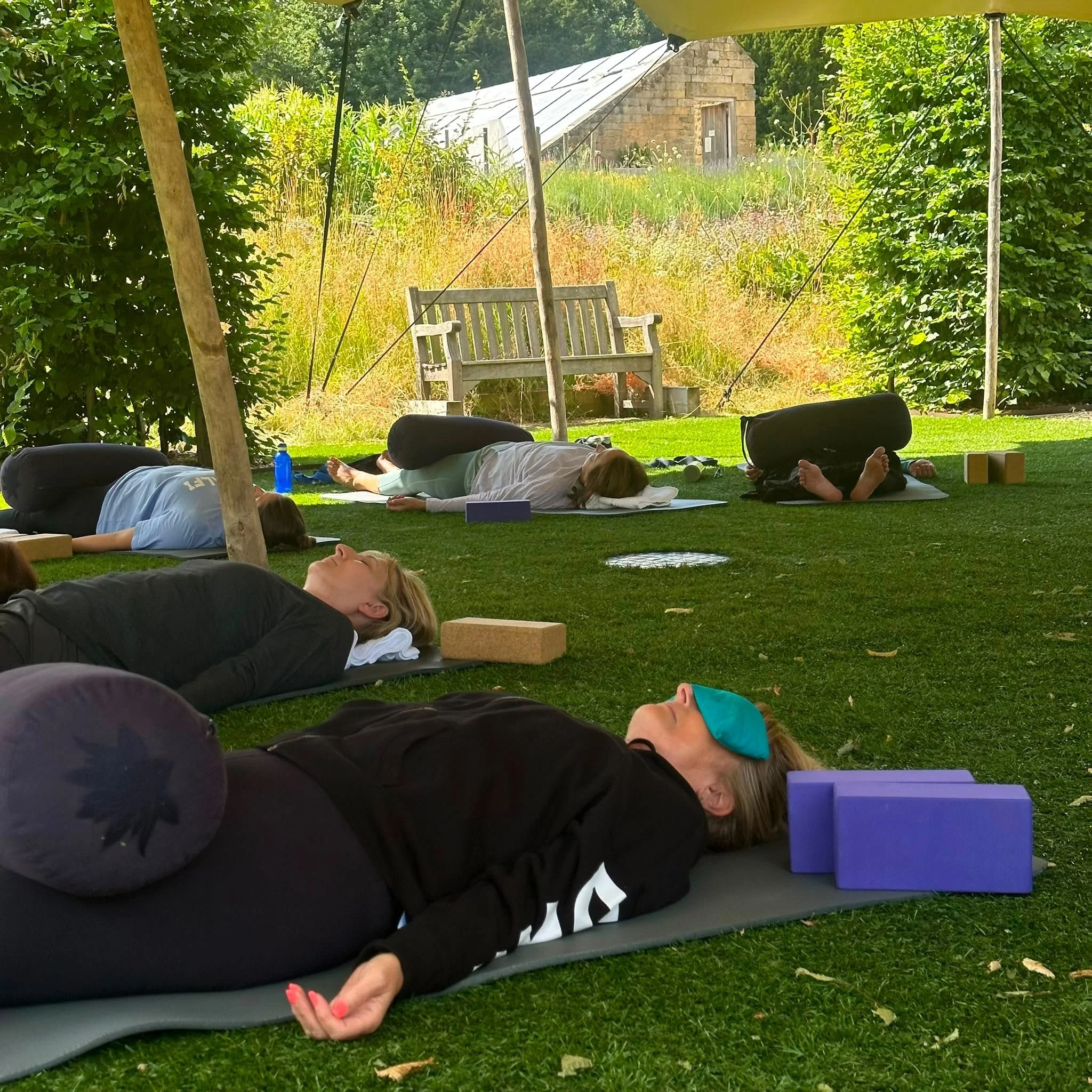 People lying on yoga mats outdoors under a canopy, practicing relaxation or meditation in a garden setting with grass, trees, and a small stone building in the background.
