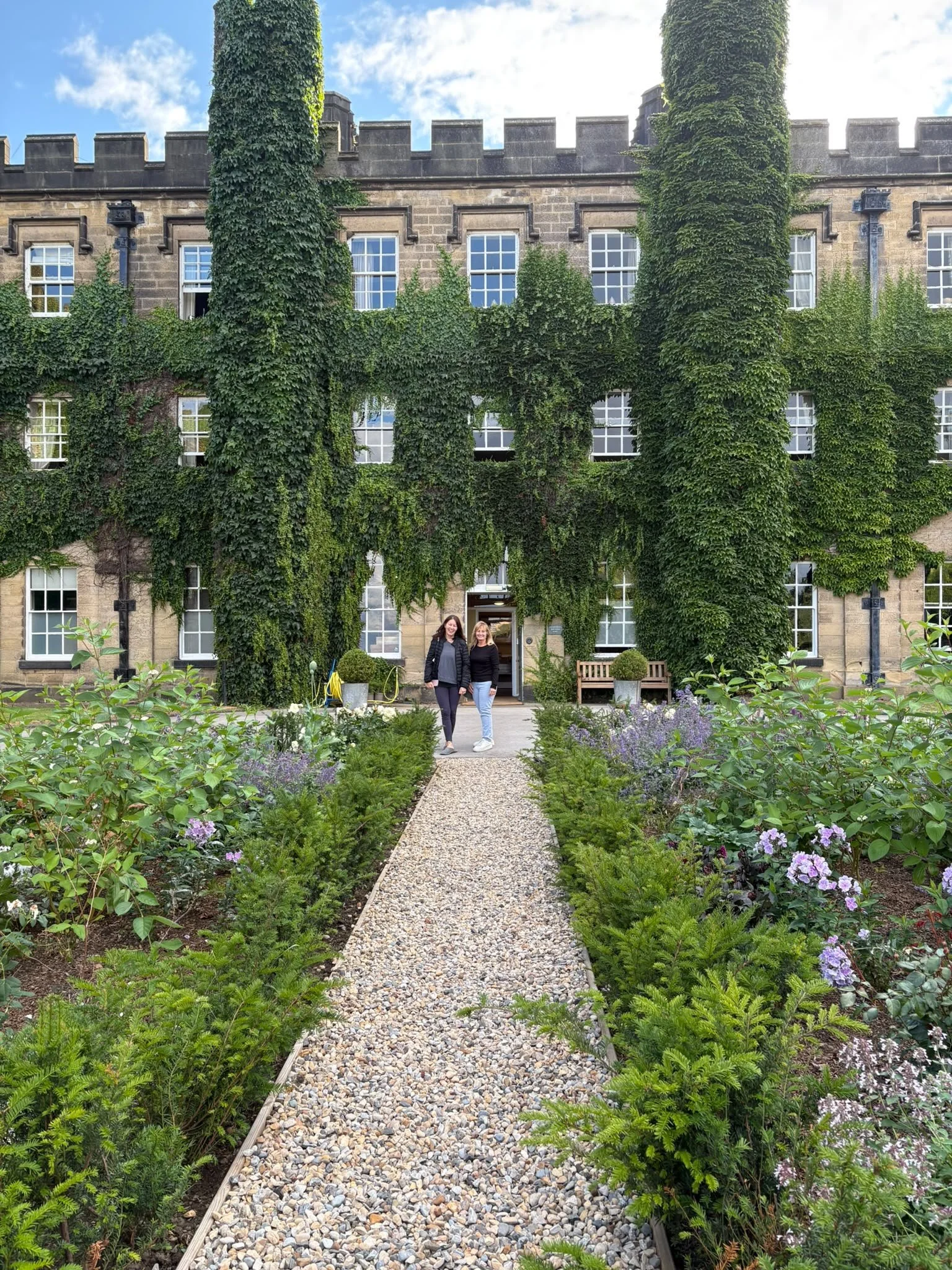 Two women standing on a gravel pathway in a garden in front of an ivy-covered brick building with multiple windows.
