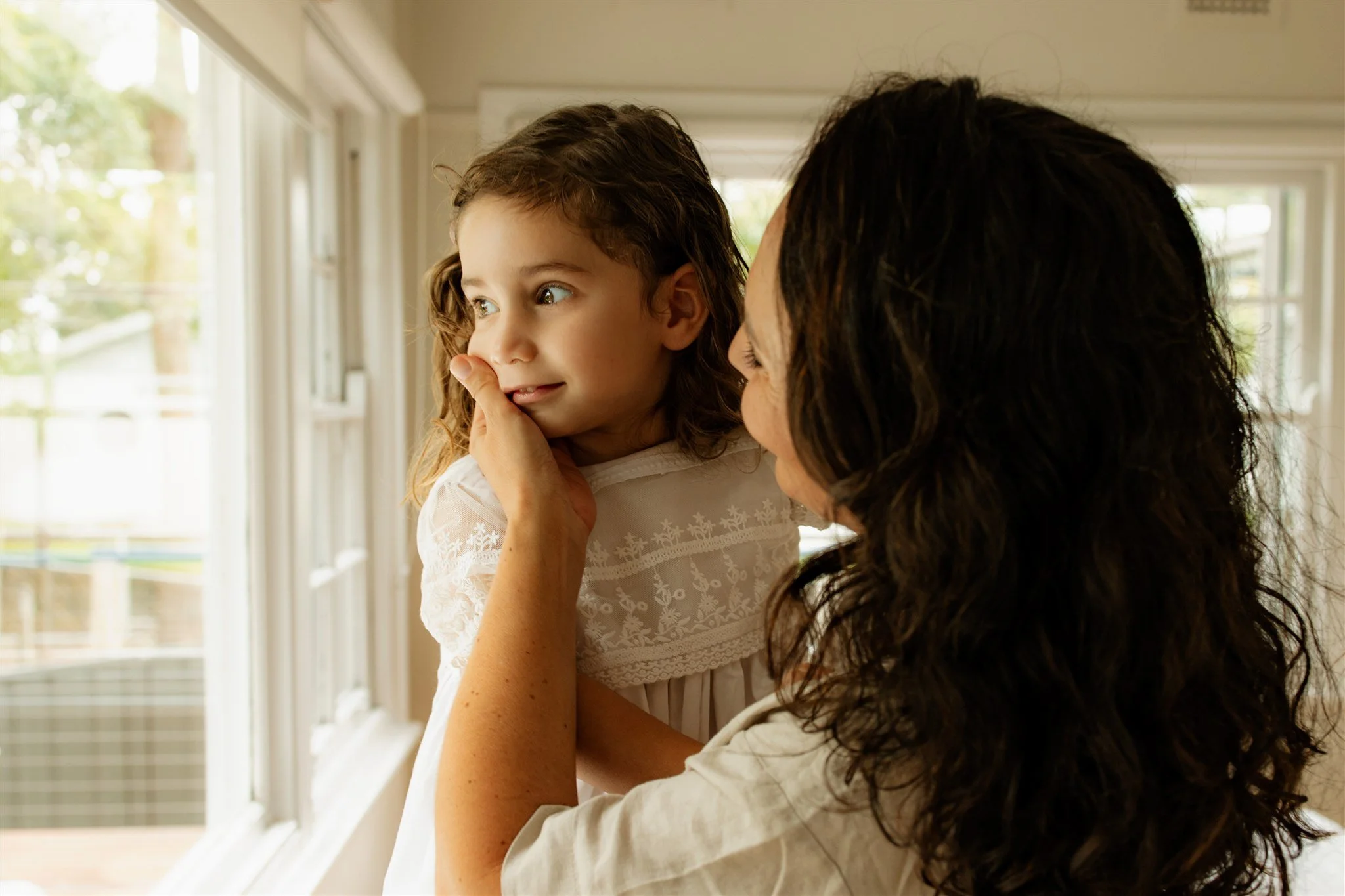 A woman holding a young girl near a window, the girl has her finger on her lips and looks to the side.