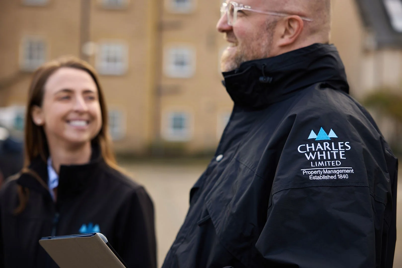 Two people outdoors wearing black jackets with 'Charles White Limited Property Management Established 1840' logo, smiling and talking.