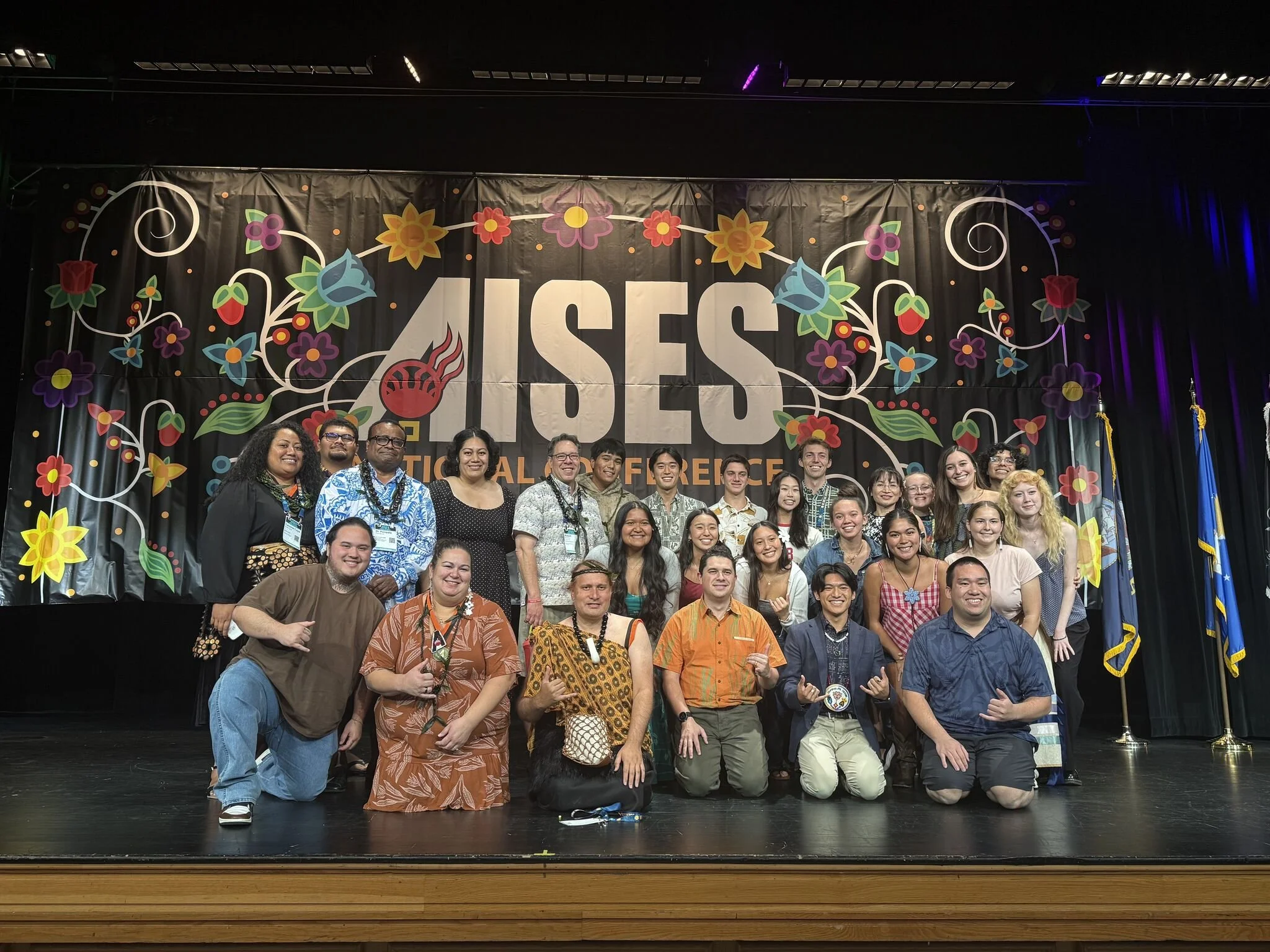 Group of people on stage at a conference with a large banner reading 'AISES' and colorful floral decorations.