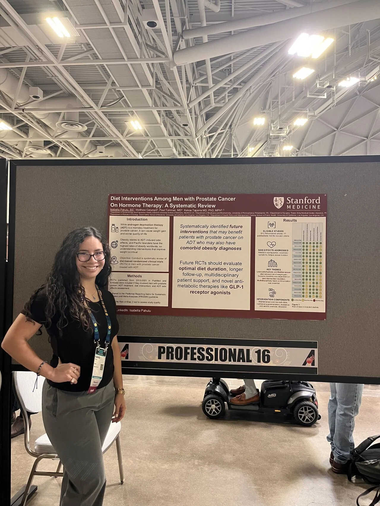 A woman with long curly hair wearing glasses, a black blouse, and gray pants is standing next to a presentation poster at a professional conference. The poster discusses diet interventions among men with prostate cancer and hormone therapy, and is affiliated with Stanford Medicine. The setting appears to be a large indoor conference or expo hall with a high ceiling and industrial lighting.