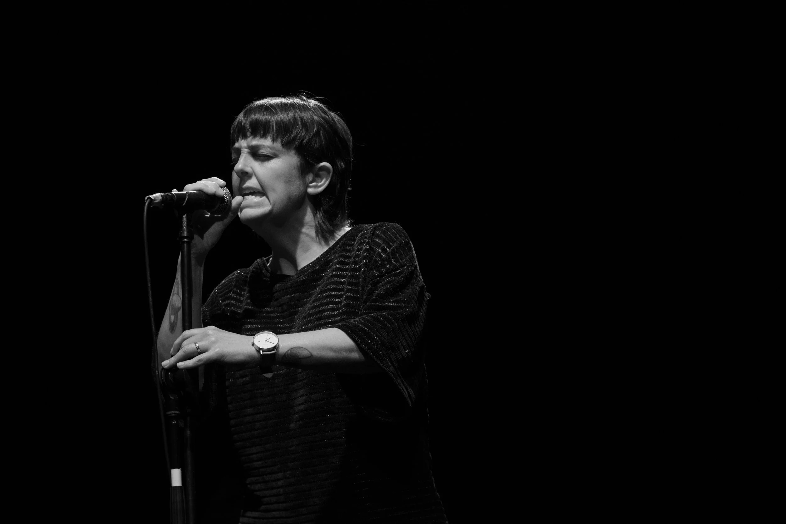 Jenny, woman with short dark brown hair and black top, holding a microphone on a stand with two hands with an intense look on her face. Eyes are closed