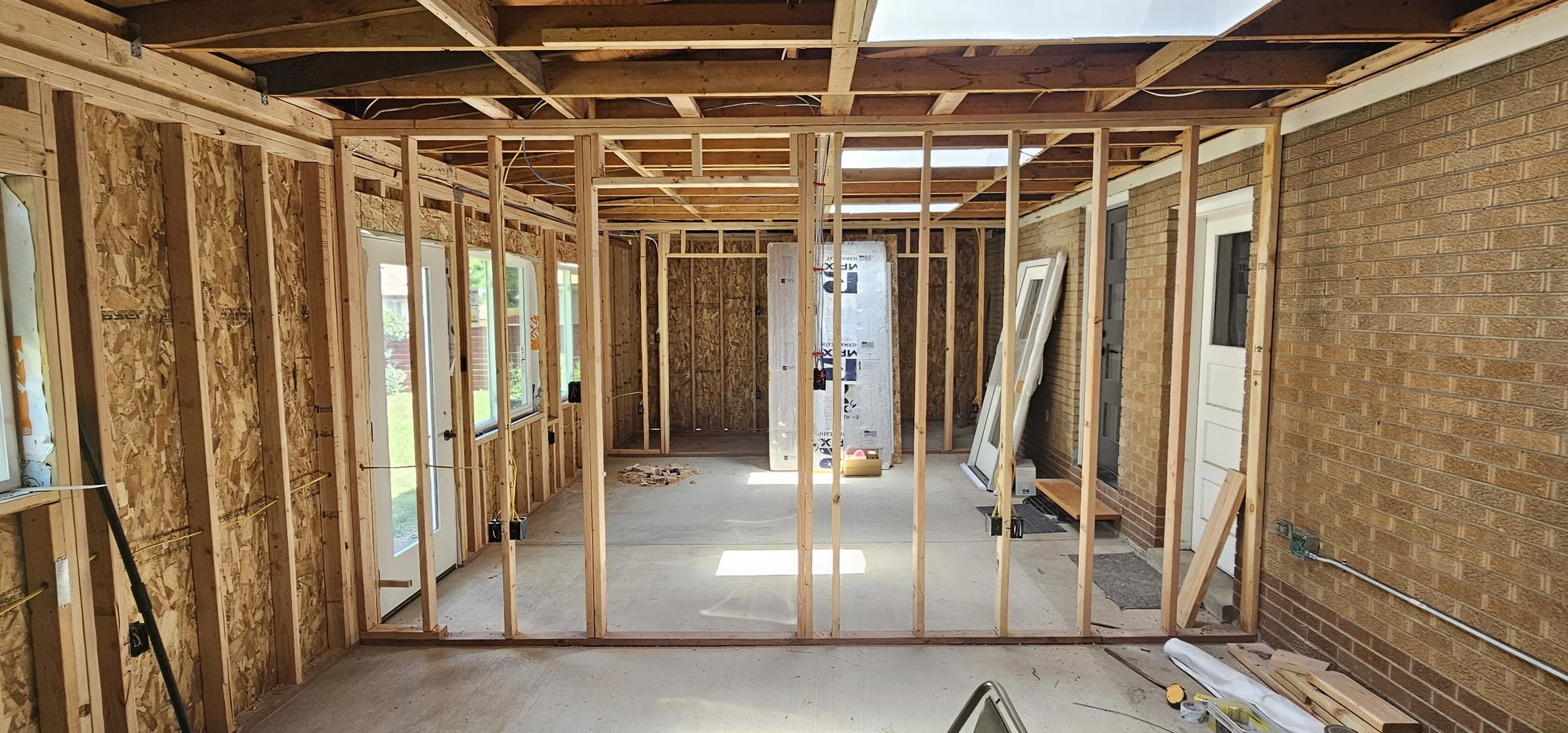 Interior of a house under construction with exposed wooden framing, OSB wall sheathing, and a brick exterior wall. There are windows and doors installed, and construction materials and tools scattered on the floor.