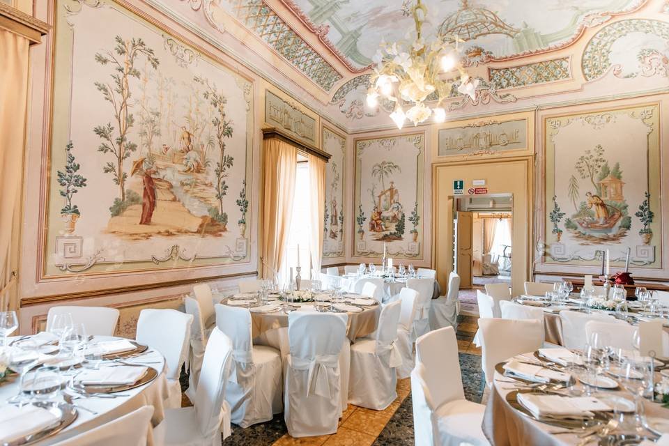 Sala da pranzo elegante con tavoli rotondi apparecchiati, sedie con copertura bianca, affreschi decorativi sulle pareti e un grande lampadario centrale.
