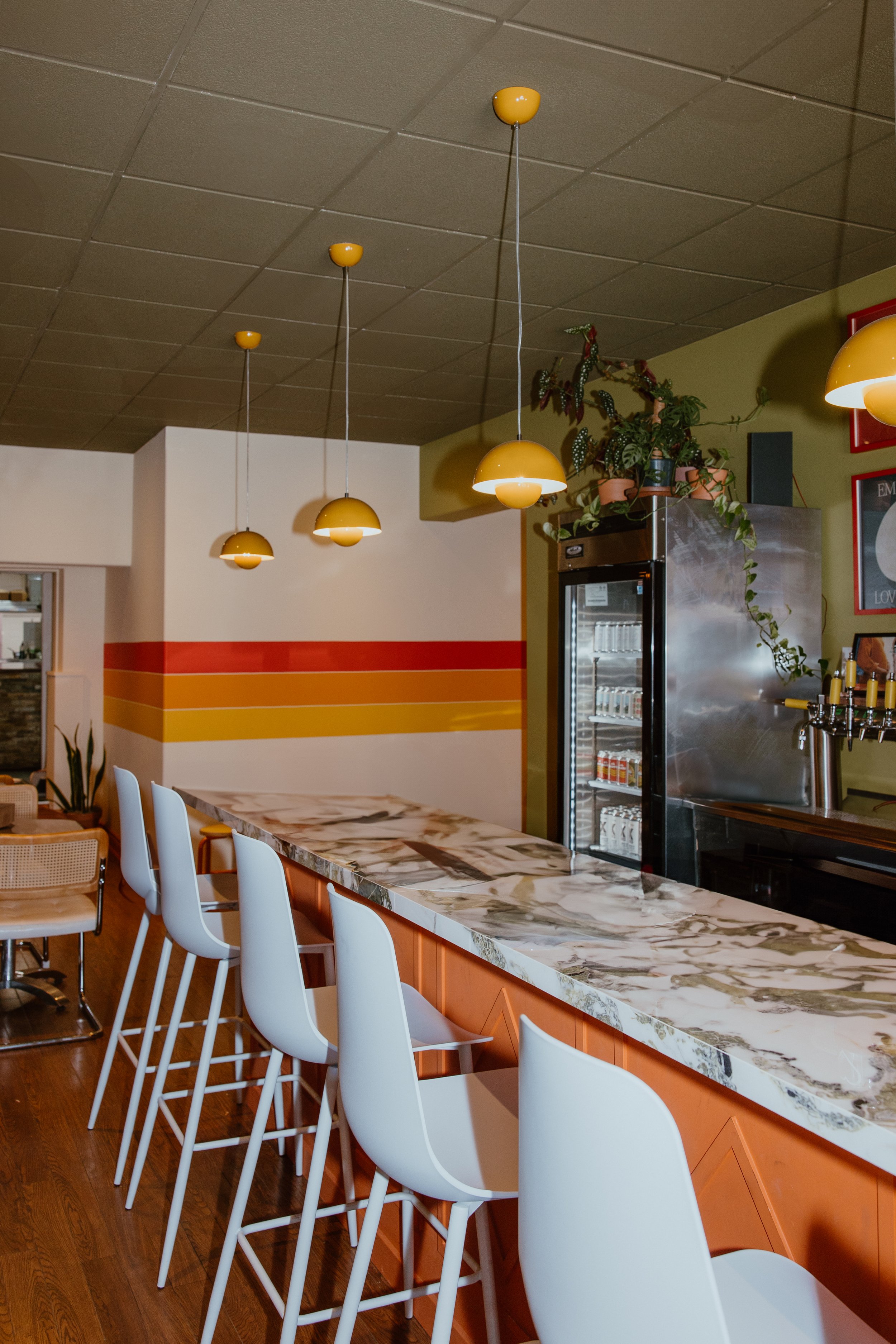 Modern restaurant bar with a marble countertop, white stools, yellow hanging pendant lights, and potted plants on top of stainless steel refrigerator.