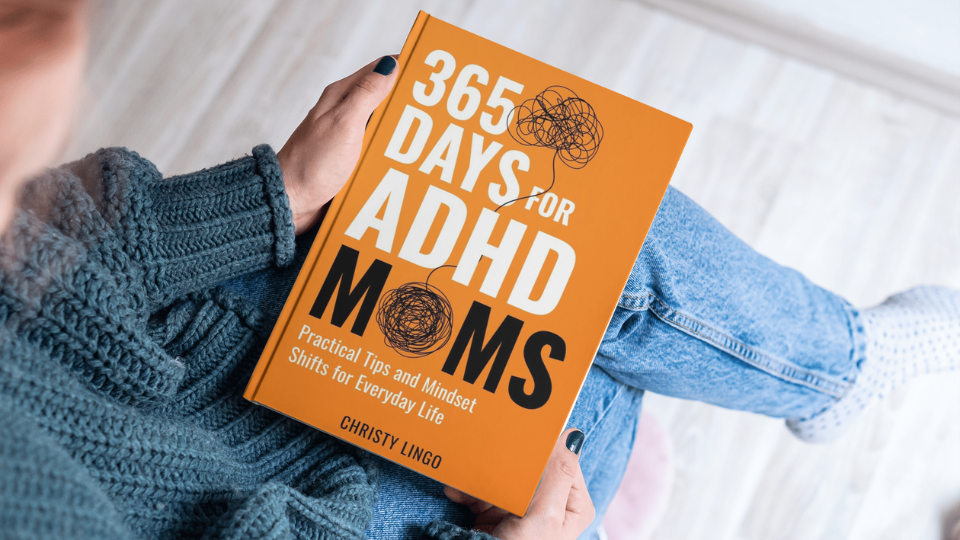 Person sitting with legs crossed reading a book titled '365 Days for ADHD Moms'  by Christy Lingo with a bright orange cover featuring a scribbled brain illustration.