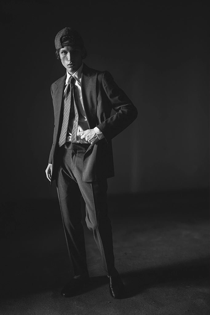 Black and white photo of a person in a suit and tie, wearing a baseball cap, standing in a dark room.