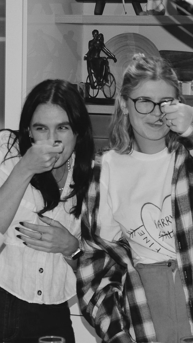 Two women are sitting side by side, both holding their noses. The woman on the left has dark hair, wears a light-colored shirt, and has her hand on her chest. The woman on the right has light hair, glasses, and wears a t-shirt with a heart and names 'HARRY' and 'SHE' written inside it, with a plaid shirt tied around her waist. They are in a room with decorative art figures, including a sculpture of a woman riding a bicycle, and other decor items in the background.