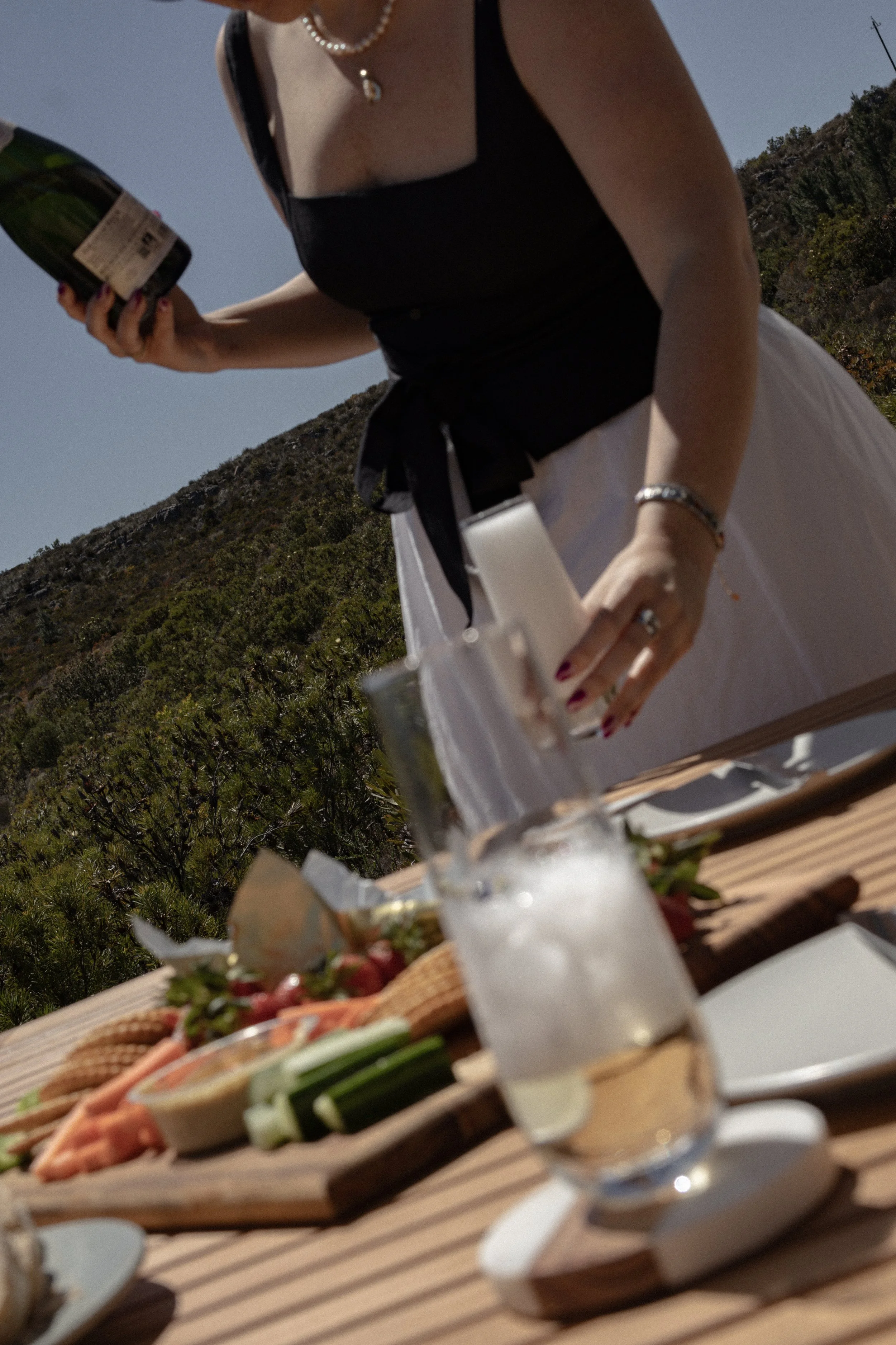 A woman wearing a black sleeveless top and white skirt, holding a wine bottle, standing outdoors with a hillside covered in shrubs and trees in the background. There is a table with food and drinks in the foreground.