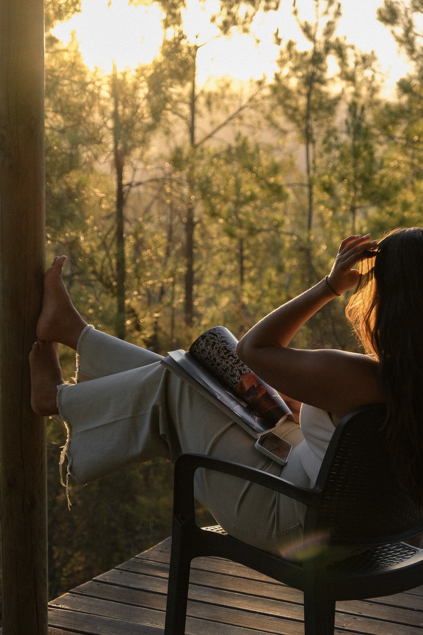 A person sitting outside on a deck, reading a magazine with a sunset view of trees in the background.
