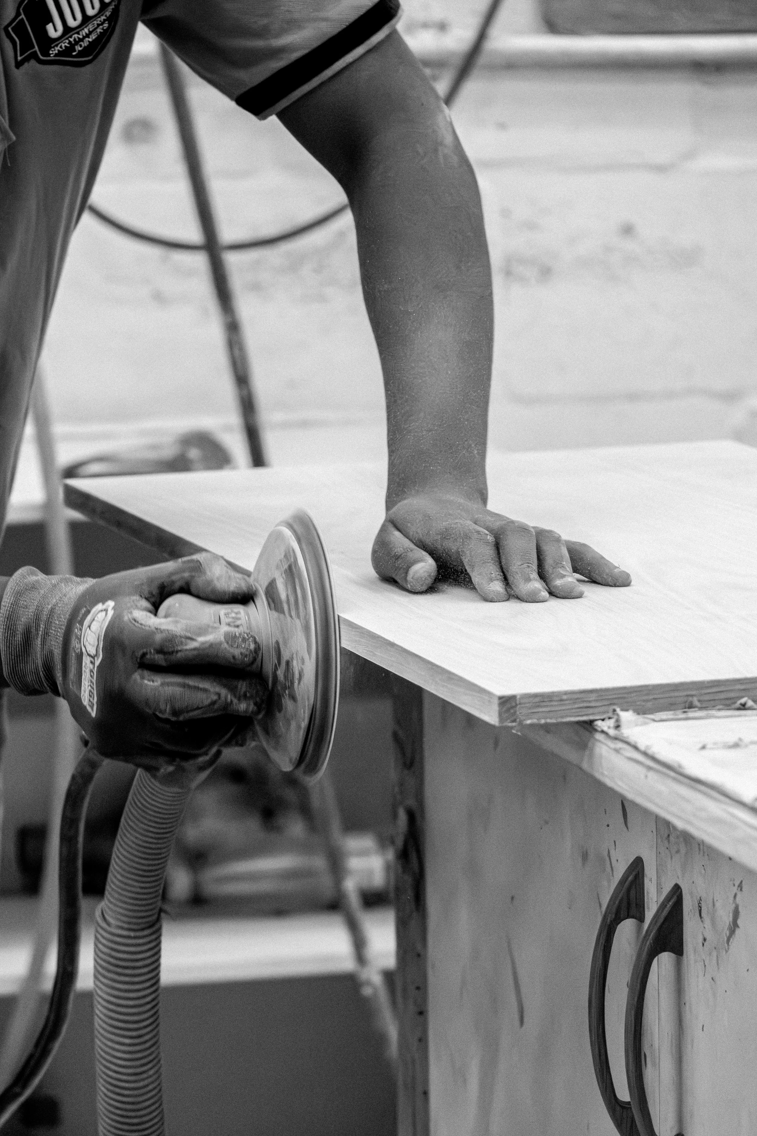 A person using an electric sander on a wooden surface, with visible dust on their hand and arm, in a workshop. The person's arm and hand are in focus, while the background shows a workbench and tools.