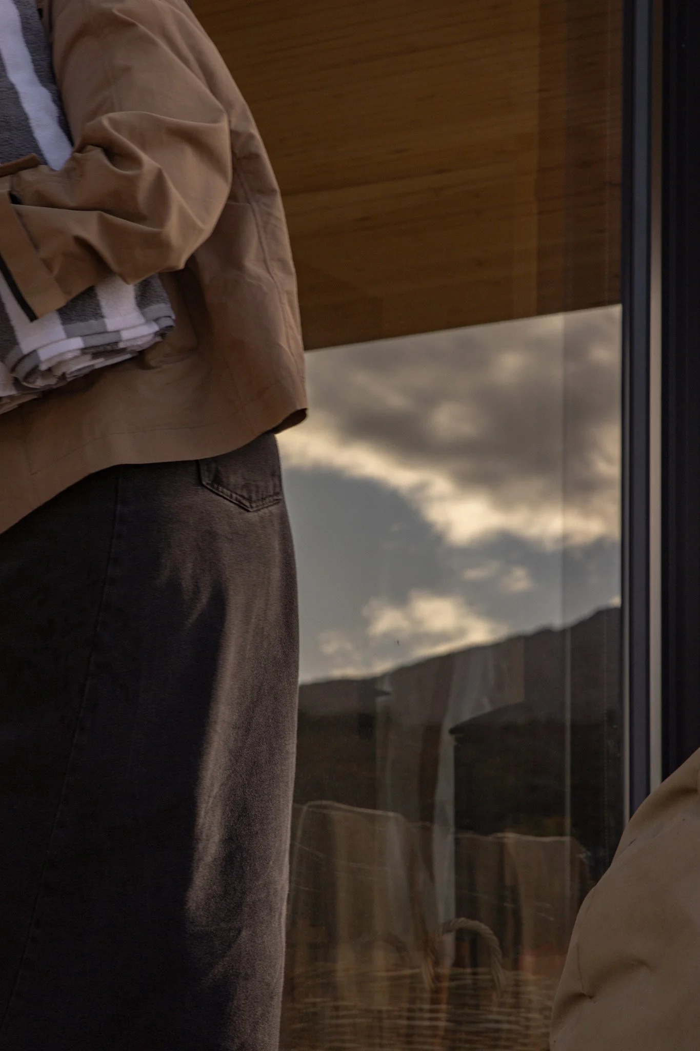 A person is lying on a bed with tan pants and a beige shirt, wrapped in a plaid blanket, in a room with wooden paneling and a large window showing cloudy skies and mountainous landscape outside.