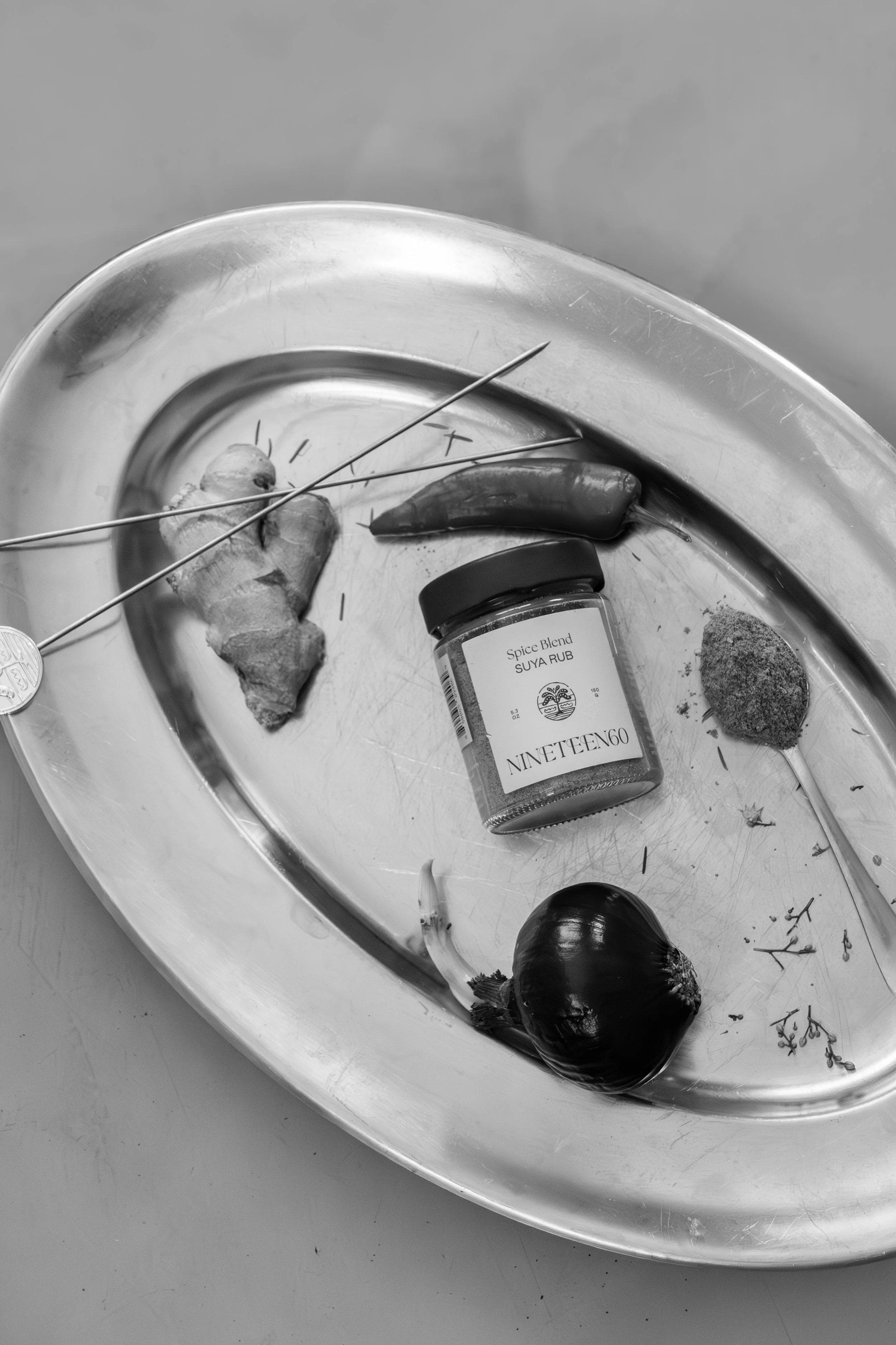 A metal tray with a ginger root, a small jar labeled 'Spice Blend SUYA RUB NINETEENGO,' a single chili pepper, an onion, and some scattered herbs and spices.