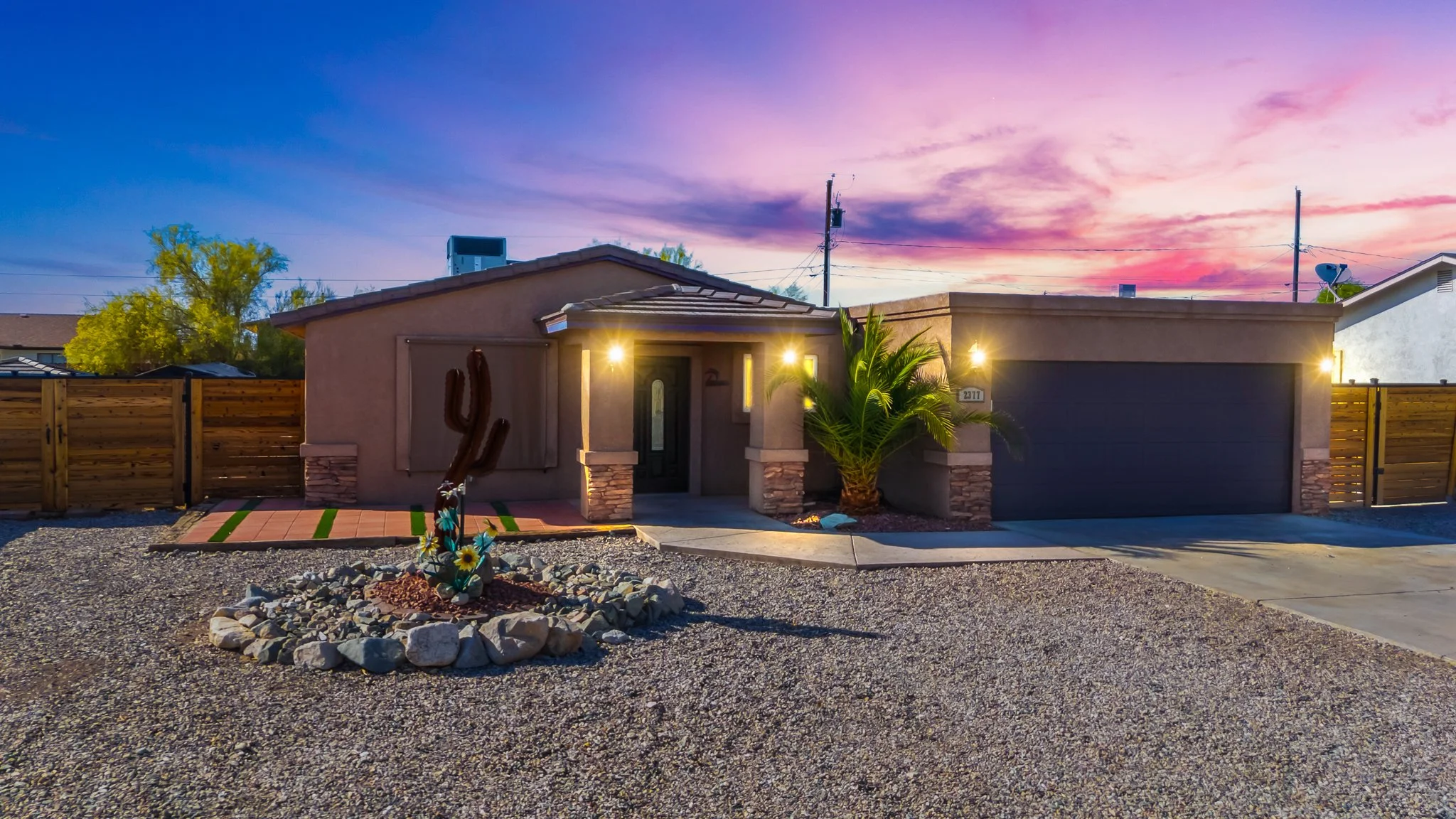 A single-story house with a gray exterior, black garage door, and a front yard featuring gravel and desert plants, set against a colorful sunset sky.