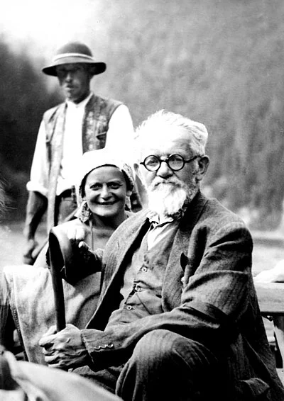 An elderly man with white hair and beard wearing glasses and a suit, sitting outdoors, with a woman and a young man standing behind him.
