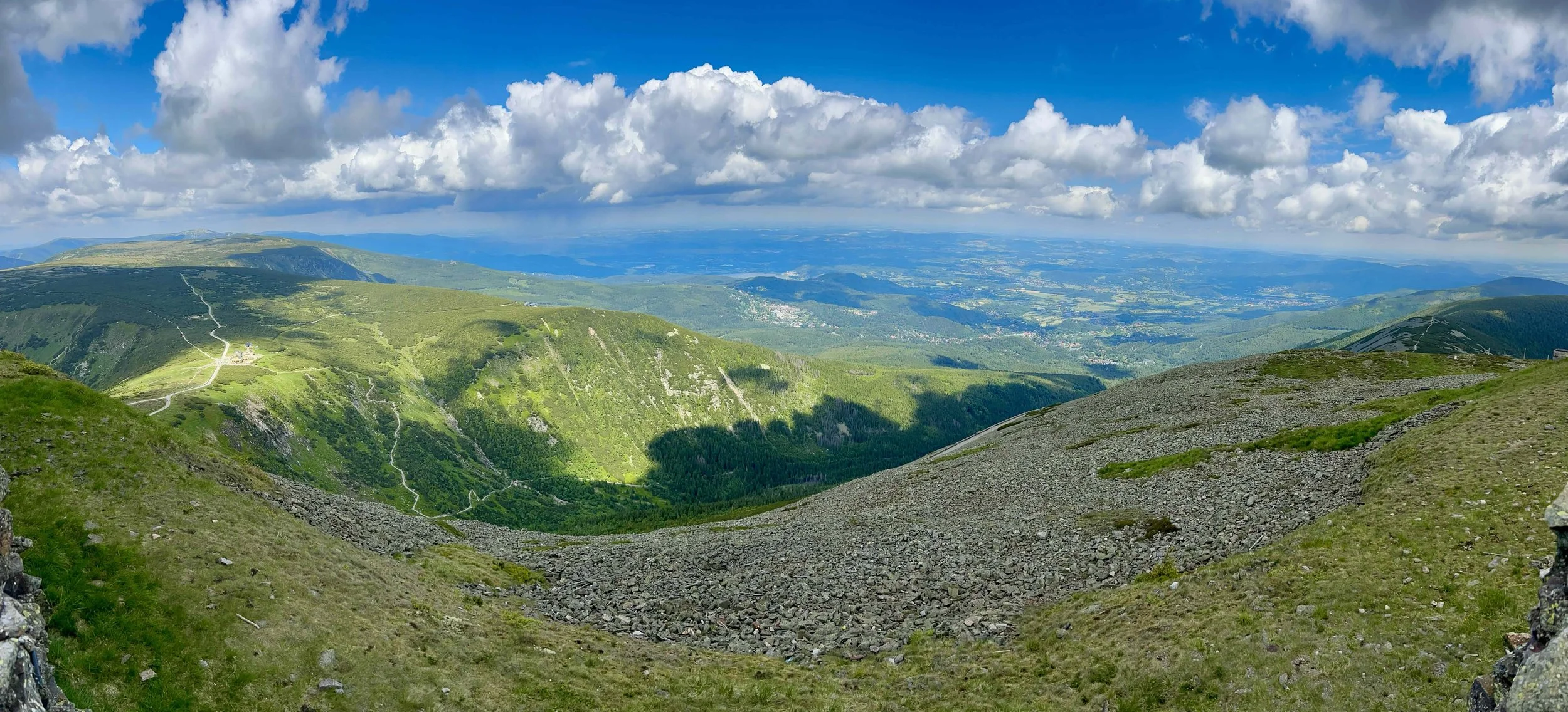 Scenic mountain landscape with green rolling hills, winding paths, and a bright blue sky with scattered clouds.