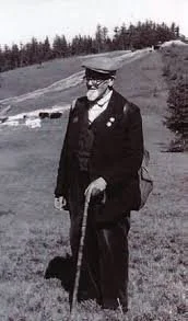 Black and white photo of a man in a military uniform with a hat and walking cane, standing outdoors on a grassy field with trees in the background.