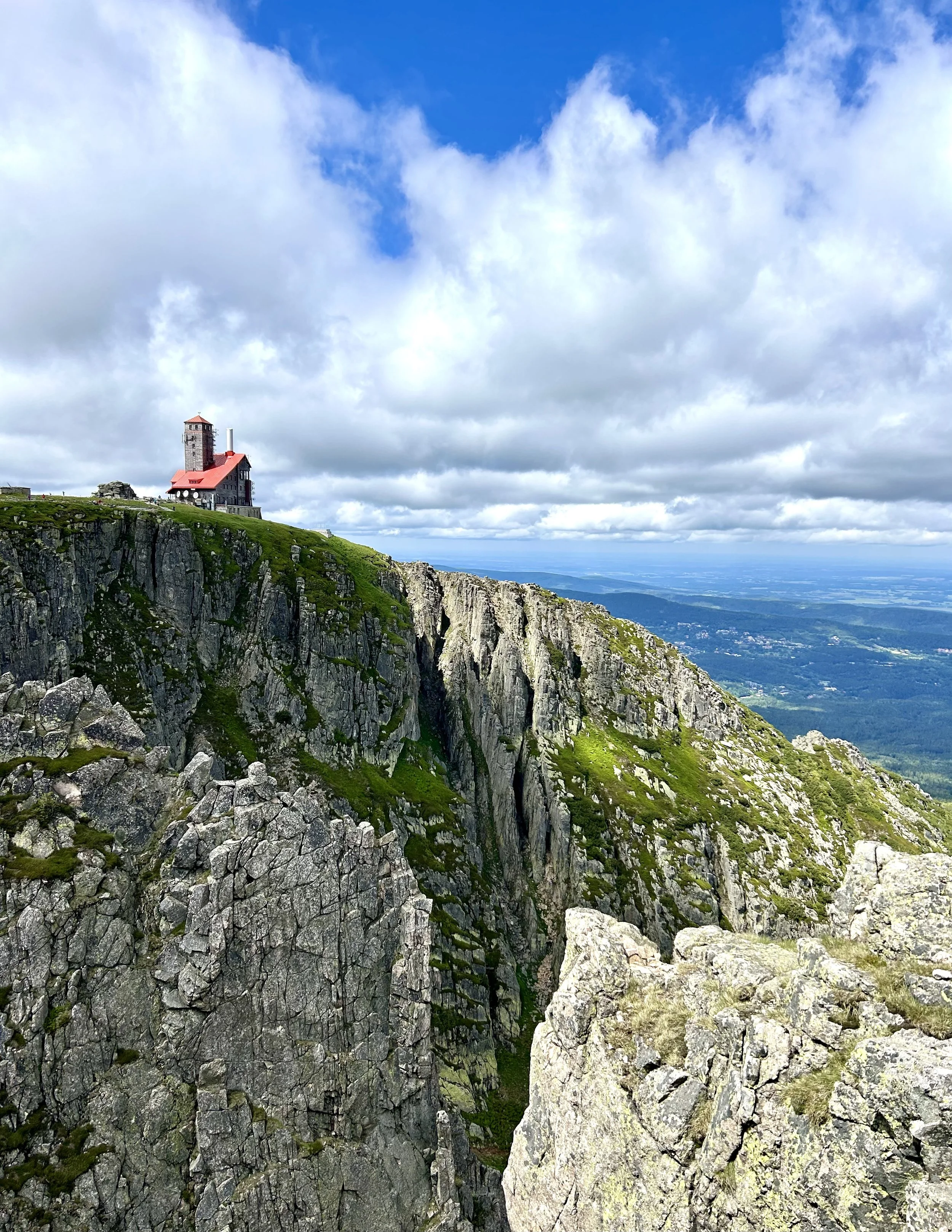 A mountain landscape with a church on a green hilltop, rugged cliffs, and a partly cloudy sky.
