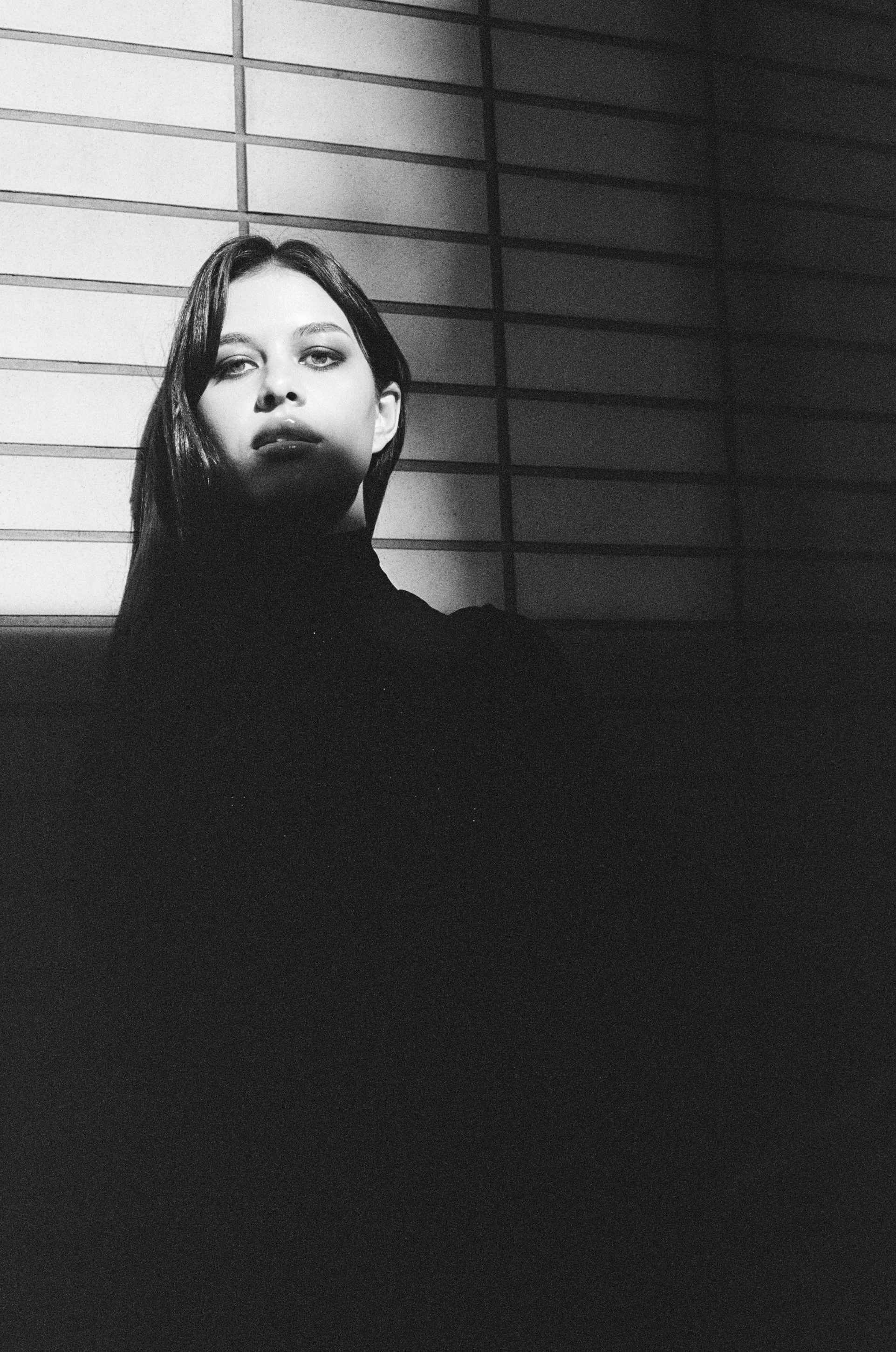 A woman with dark hair and makeup looking towards the camera, standing in front of a wall with horizontal lines, in black and white.