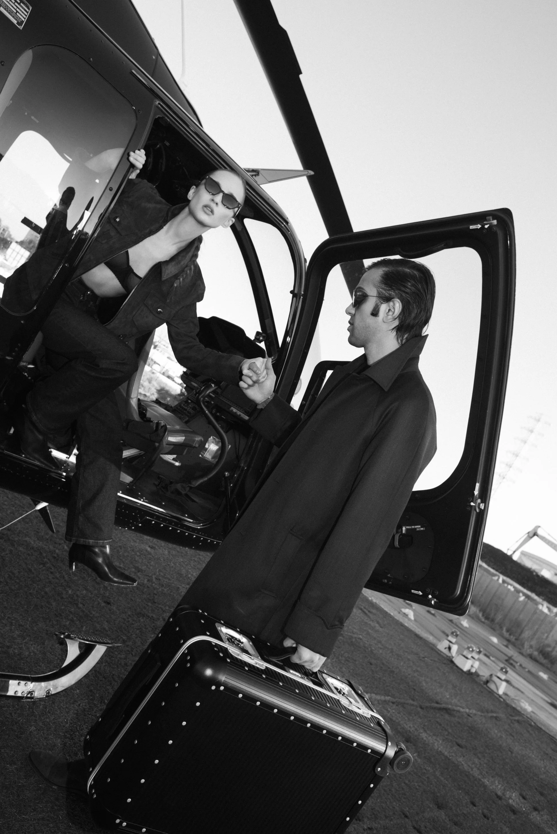 A black and white photograph of a woman in sunglasses and a dark jacket leaning out of an helicopter, shaking hands with a man wearing sunglasses and a dark coat, standing outside the helicopter in an open area with a clear sky.