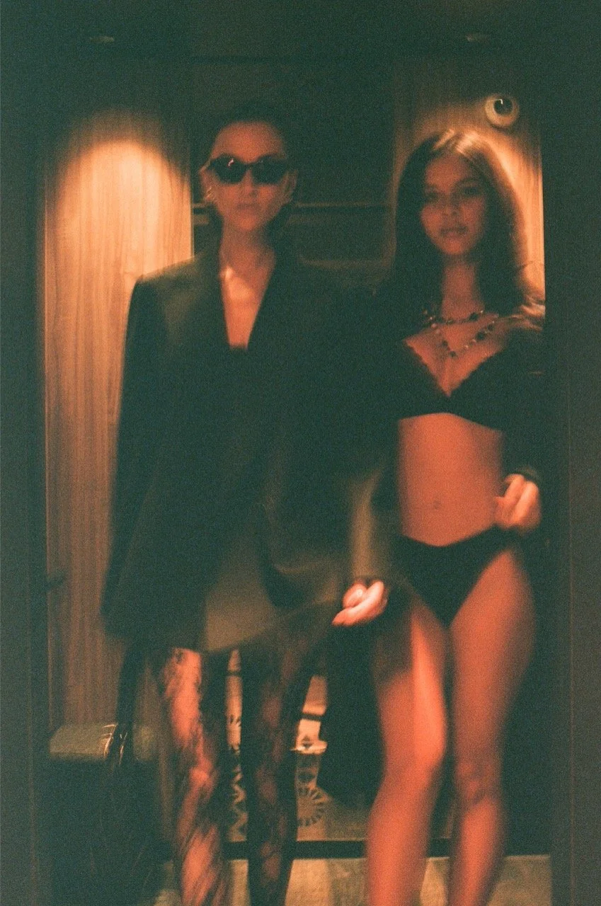 Two women standing in a dimly lit room with wood paneling. One woman is dressed in dark clothing with sunglasses, and the other is in black lingerie with jewelry.