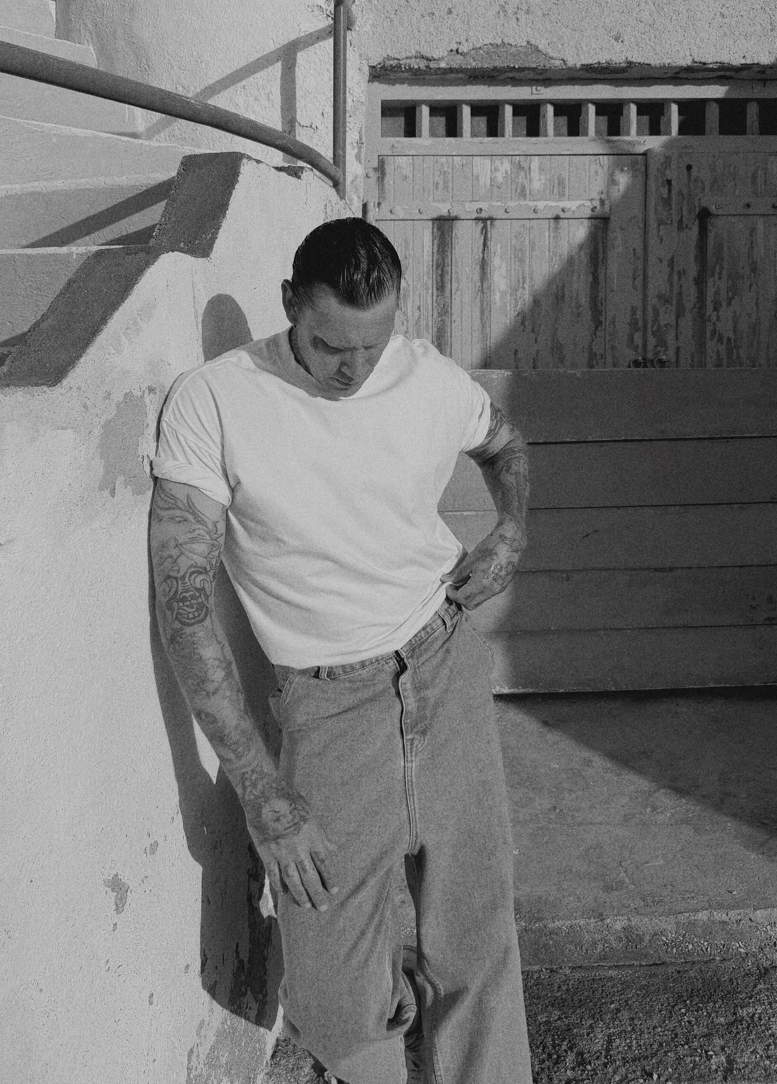 A man with tattoos on his arms, wearing a white t-shirt, stands against a wall with one arm bent and looking downward, with his hand inside his pants pocket, in front of a wooden gate or door.