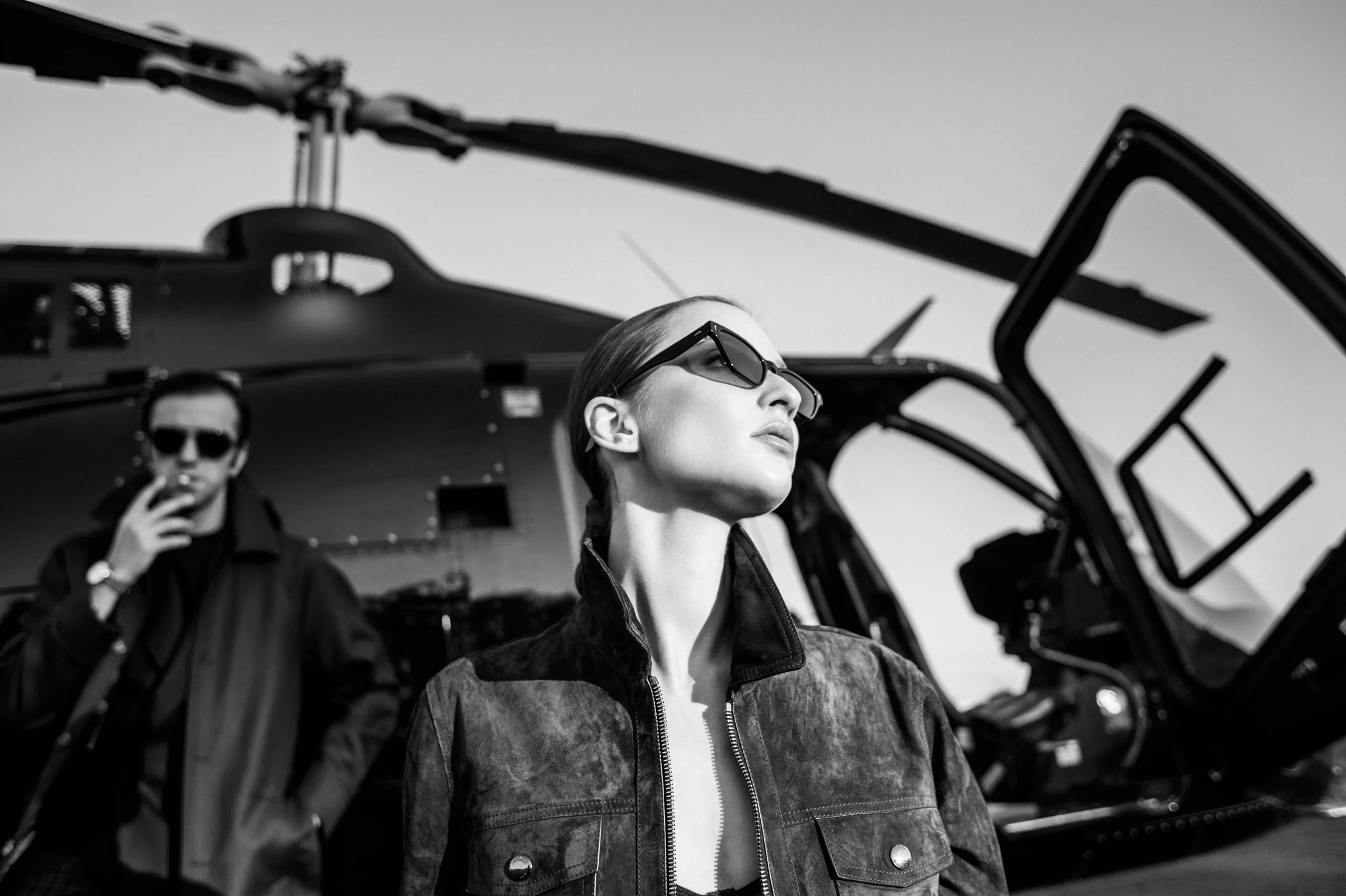 Two women with sunglasses standing near helicopters, one in the foreground and one in the background, on a clear day.