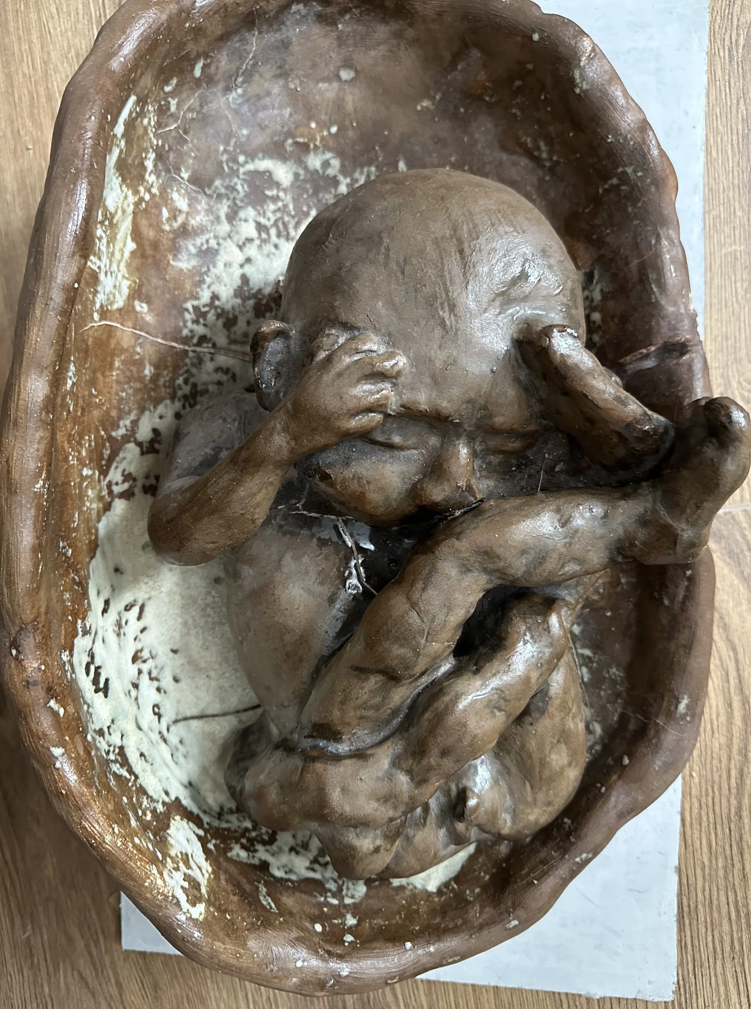 A sculpture of a baby with closed eyes, holding its head with one hand, lying in a wooden cradle.