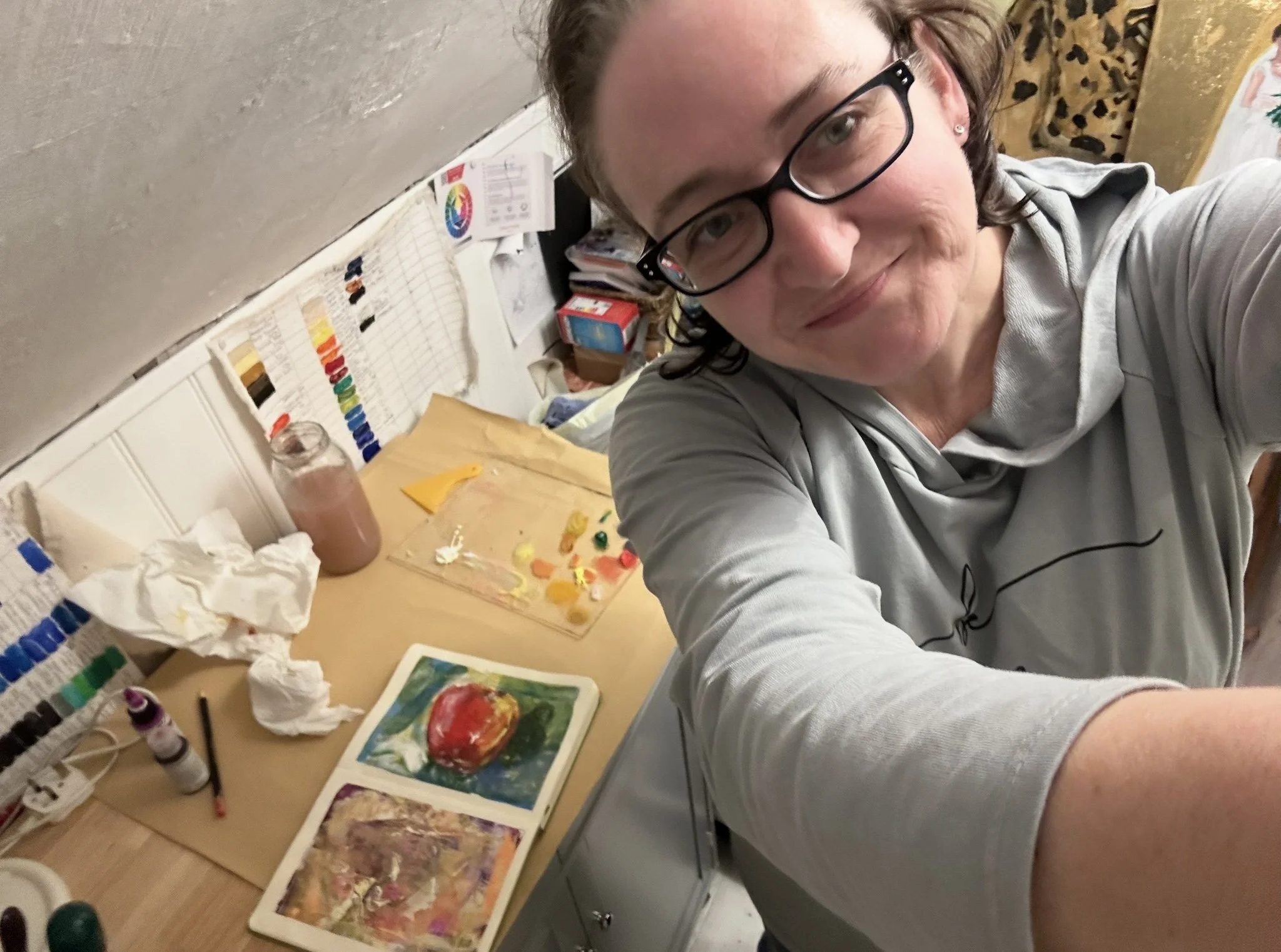 A woman in glasses taking a selfie in her art studio, with paintings, paints, brushes, and art supplies on the table behind her.