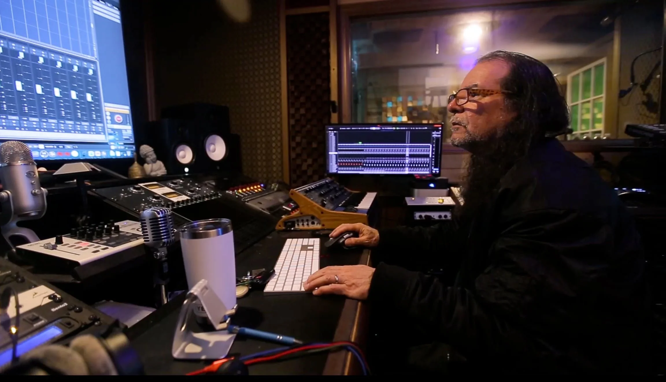 A person with long dark hair and glasses working at a music studio surrounded by audio equipment, computers, and microphones.