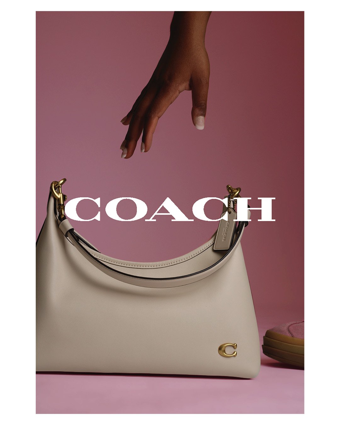 A fashion advertisement displaying a beige Coach handbag with a gold logo, set against a pink background. A hand is reaching from above, pointing toward the bag, which is partially obscured by the Coach logo text.