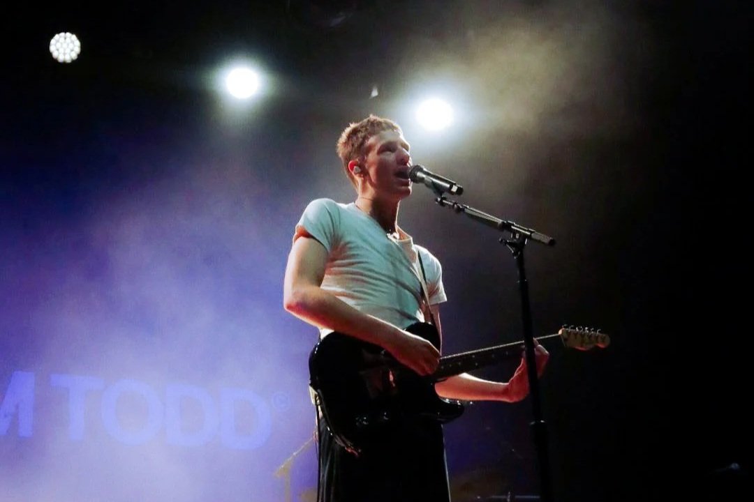 A woman with short hair singing into a microphone while playing an electric guitar on stage, illuminated by stage lights.