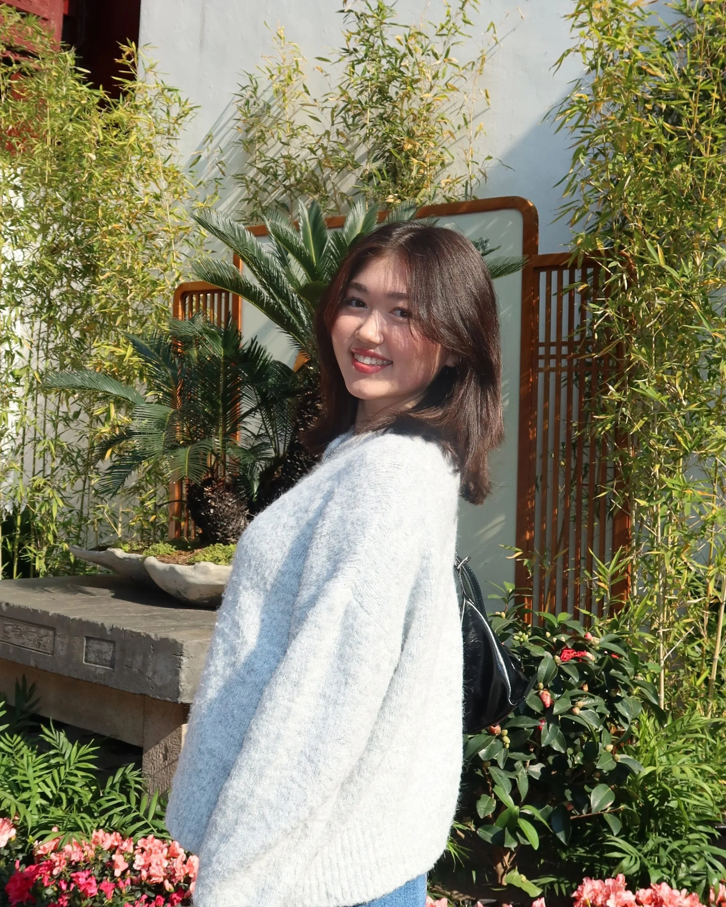 A young woman with shoulder-length brown hair smiling outdoors among green plants and pink flowers, wearing a light gray sweater and carrying a black backpack.