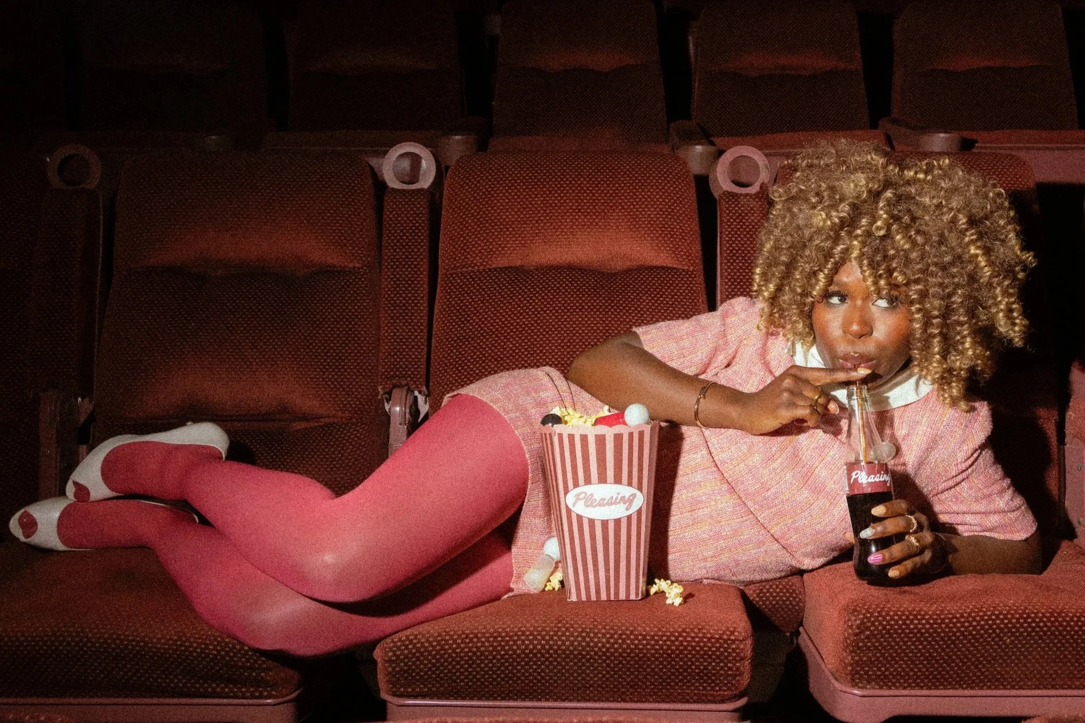 A woman with curly blonde hair lying on a row of empty red theater seats, wearing a pink outfit with matching tights and white shoes with red accents, drinking a soda from a straw.