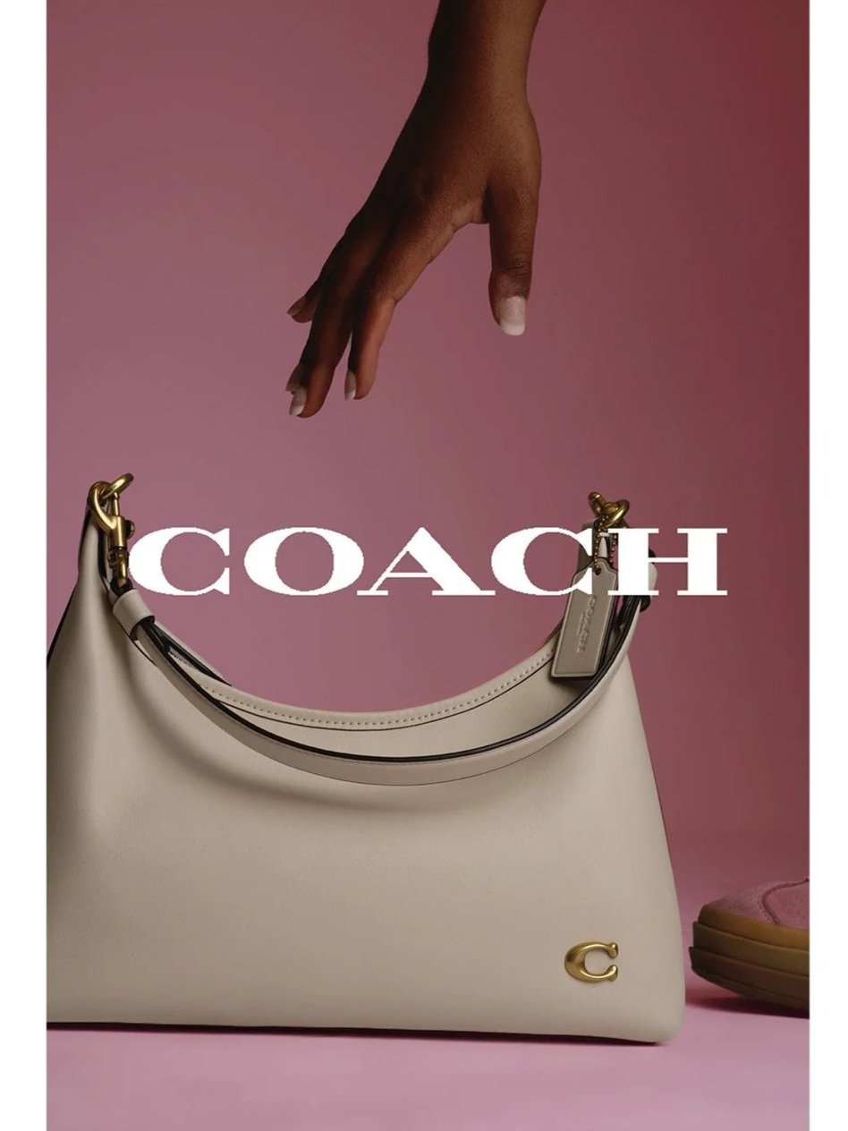 A beige Coach handbag with gold accents and a small gold 'C' logo on the lower right corner, set against a pink background, with a hand reaching from above.