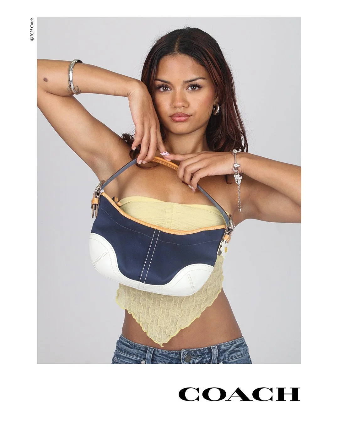A woman with shoulder-length brown hair wearing a yellow top and denim shorts holds a navy and white Coach handbag against a plain background.