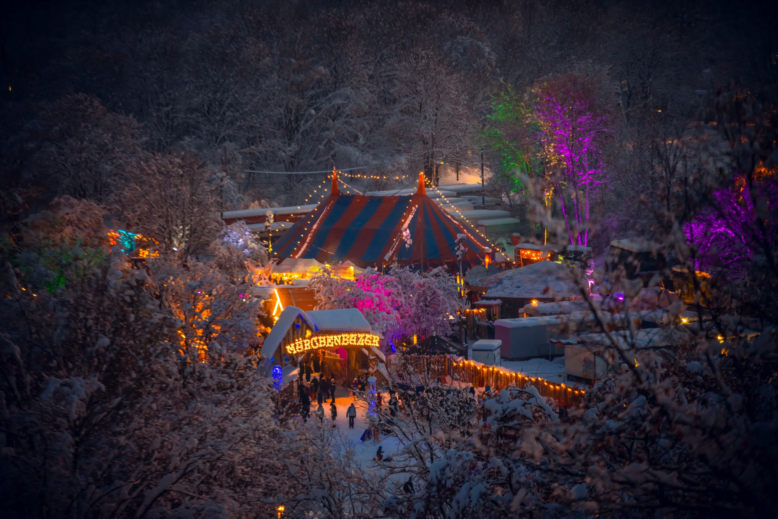 Blick auf einen festlich beleuchteten Weihnachtsmarkt im Winter, umgeben von schneebedeckten Bäumen und Zelten, mit bunten Lichtern und Menschen im Schneegarten.