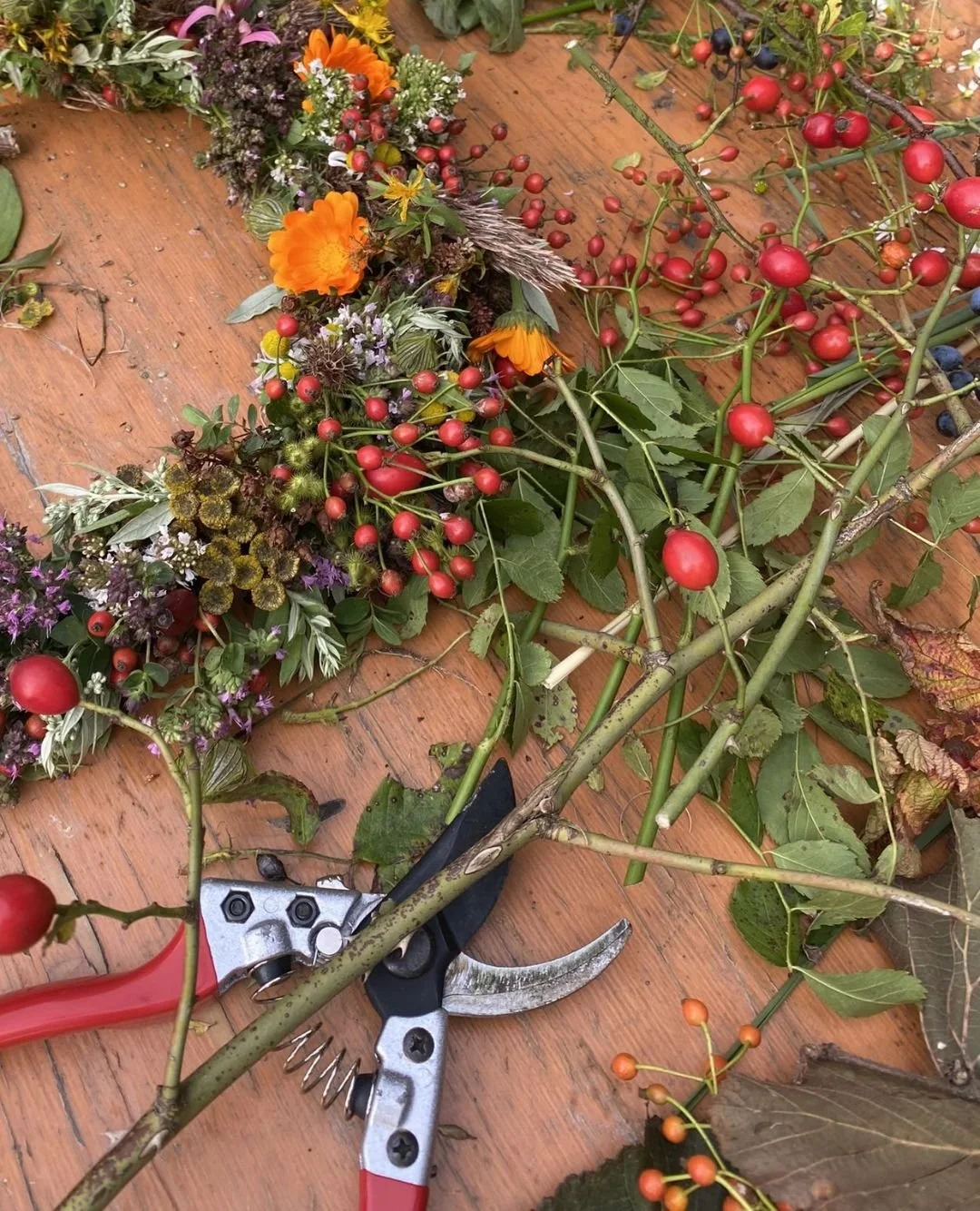 Ein Holzboden mit verschiedenen bunten Blumen, Beeren und Blättern, die für einen Blumenkranz vorbereitet werden, sowie eine Gartenschere.