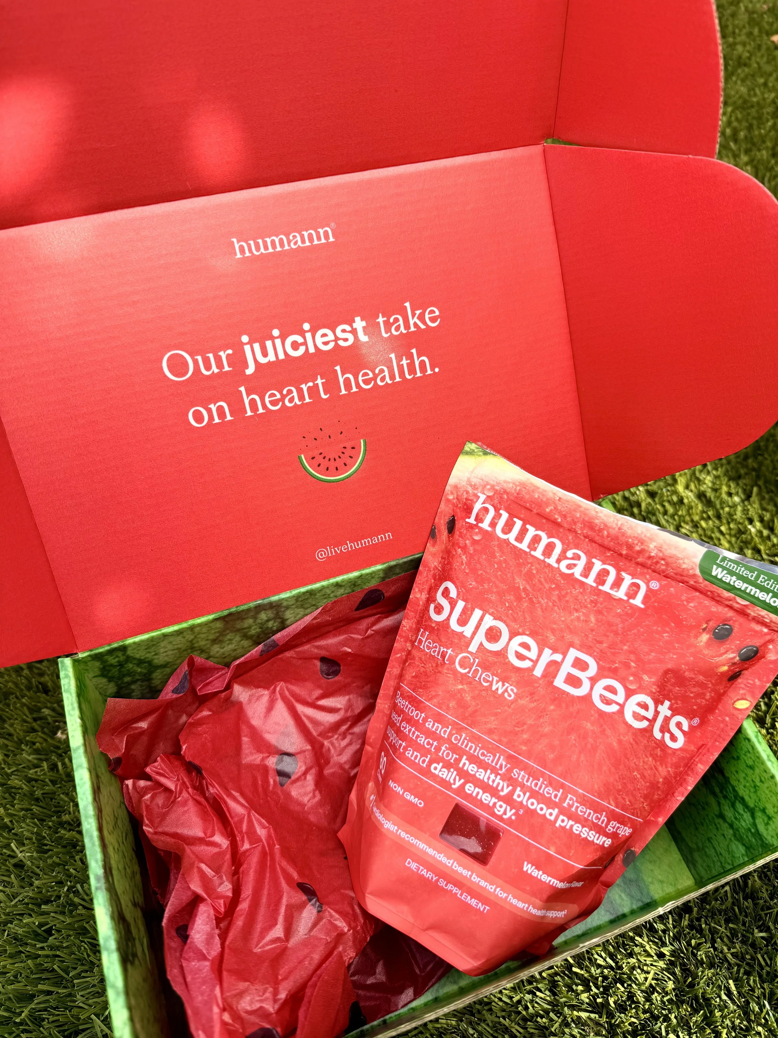 Open box of Humann SuperBeets Heart Chews with a packet inside featuring a watermelon flavor label, set on green grass with red tissue paper.