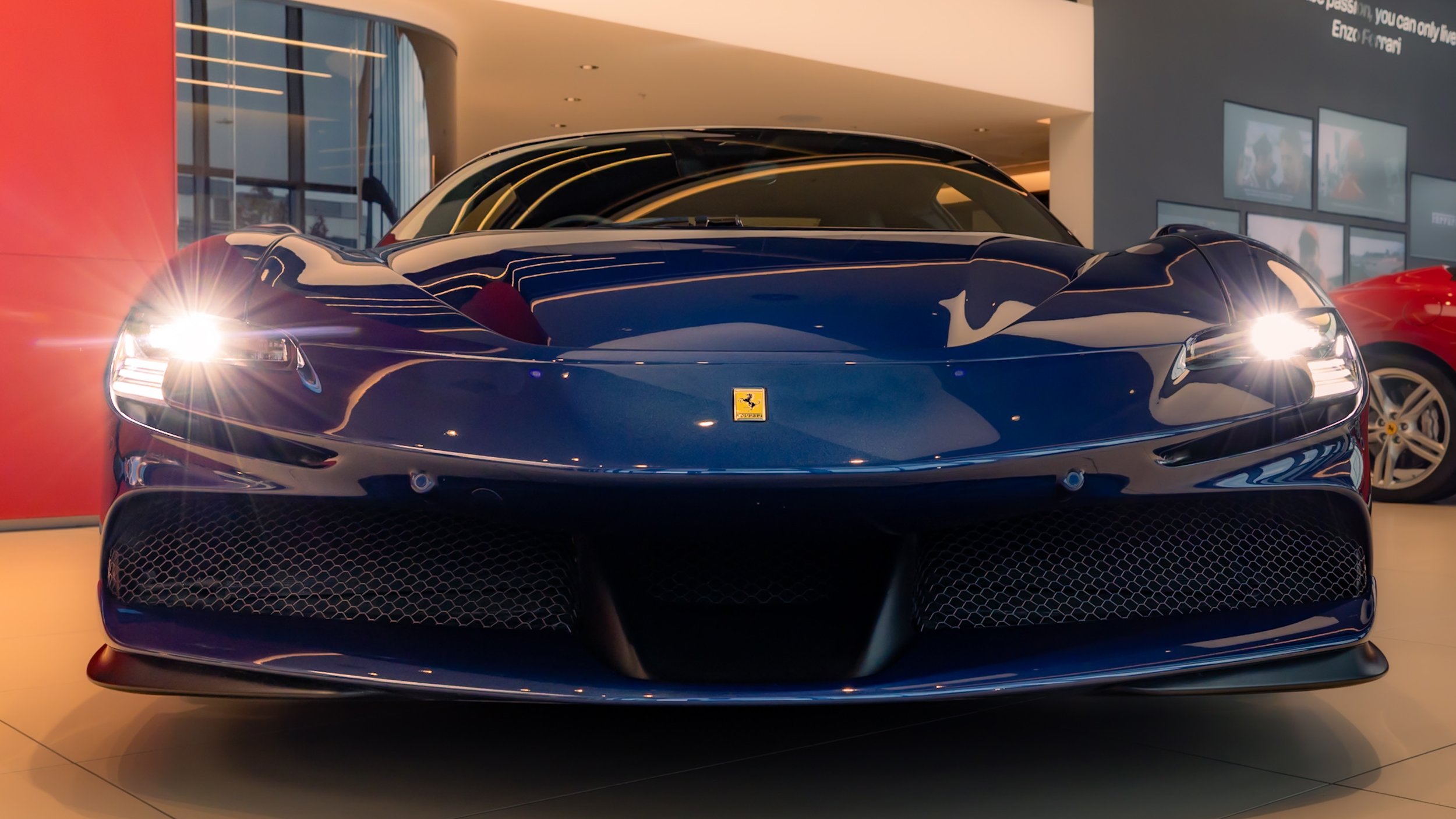 Front view of a blue sports car with headlights on, displayed indoors, showing a logo on the front bumper.